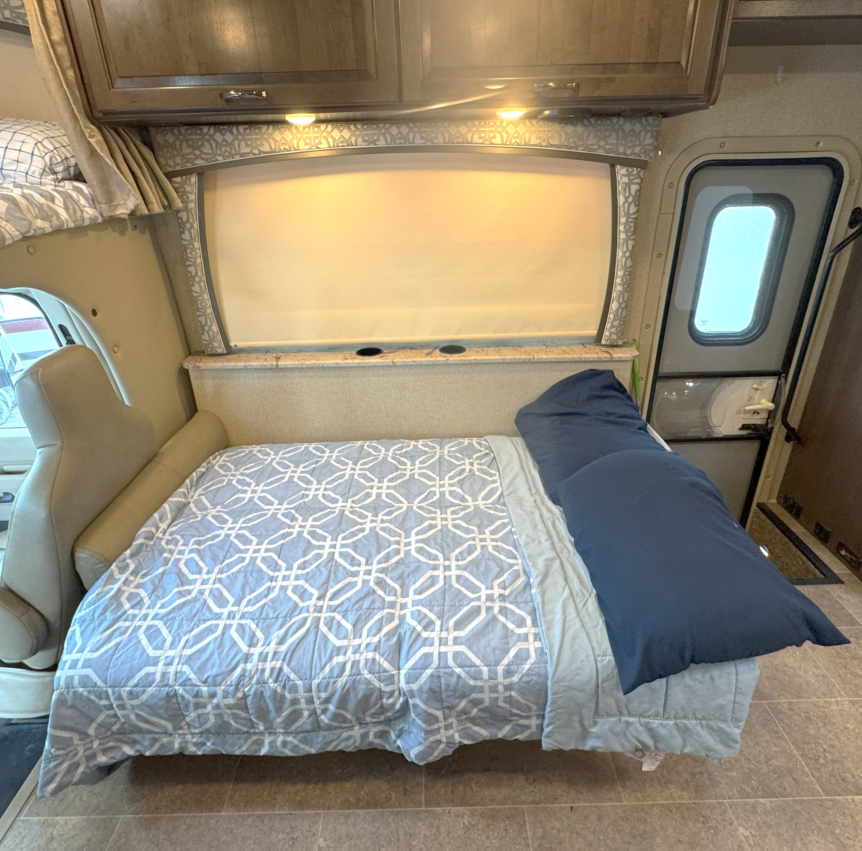 Cozy RV rental interior shows a neatly made bed with blue bedding, overhead cabinets, and passenger entry area.. Thor Chateau 2017