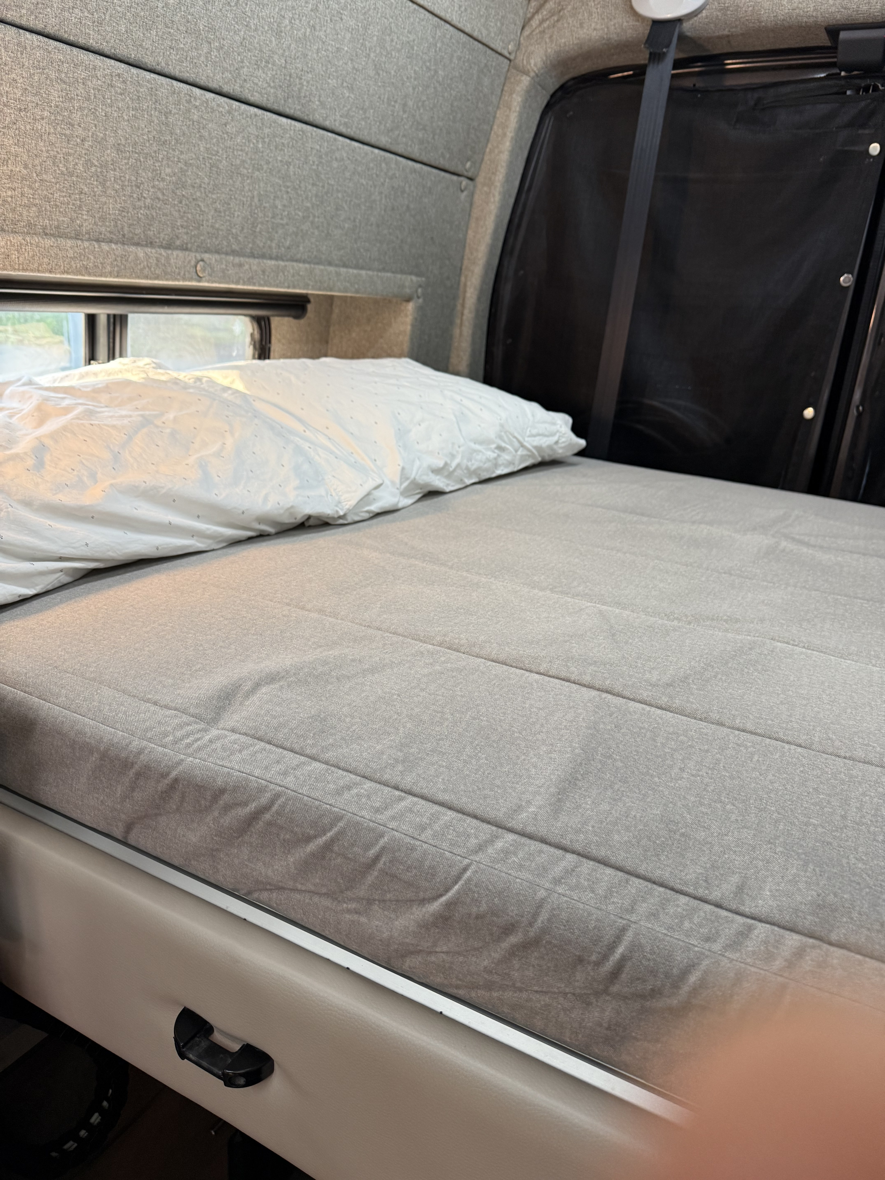 Comfortable bed area inside an RV rental, with a grey mattress, white pillow, and window view. Ready for your trip!. Adventurer Ford TRIBUTE 2021