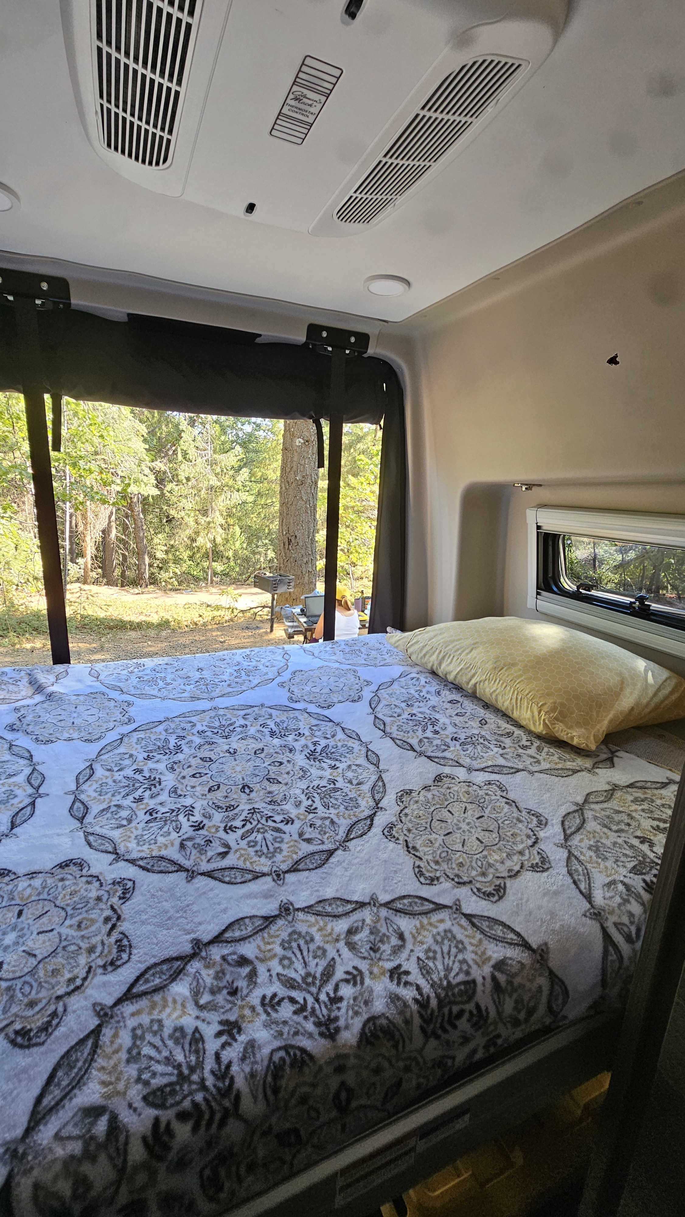 Cozy RV rental bedroom with a large window revealing a serene forest view. Features a patterned blanket, yellow pillow, and overhead AC.. Jayco Terrain 2024