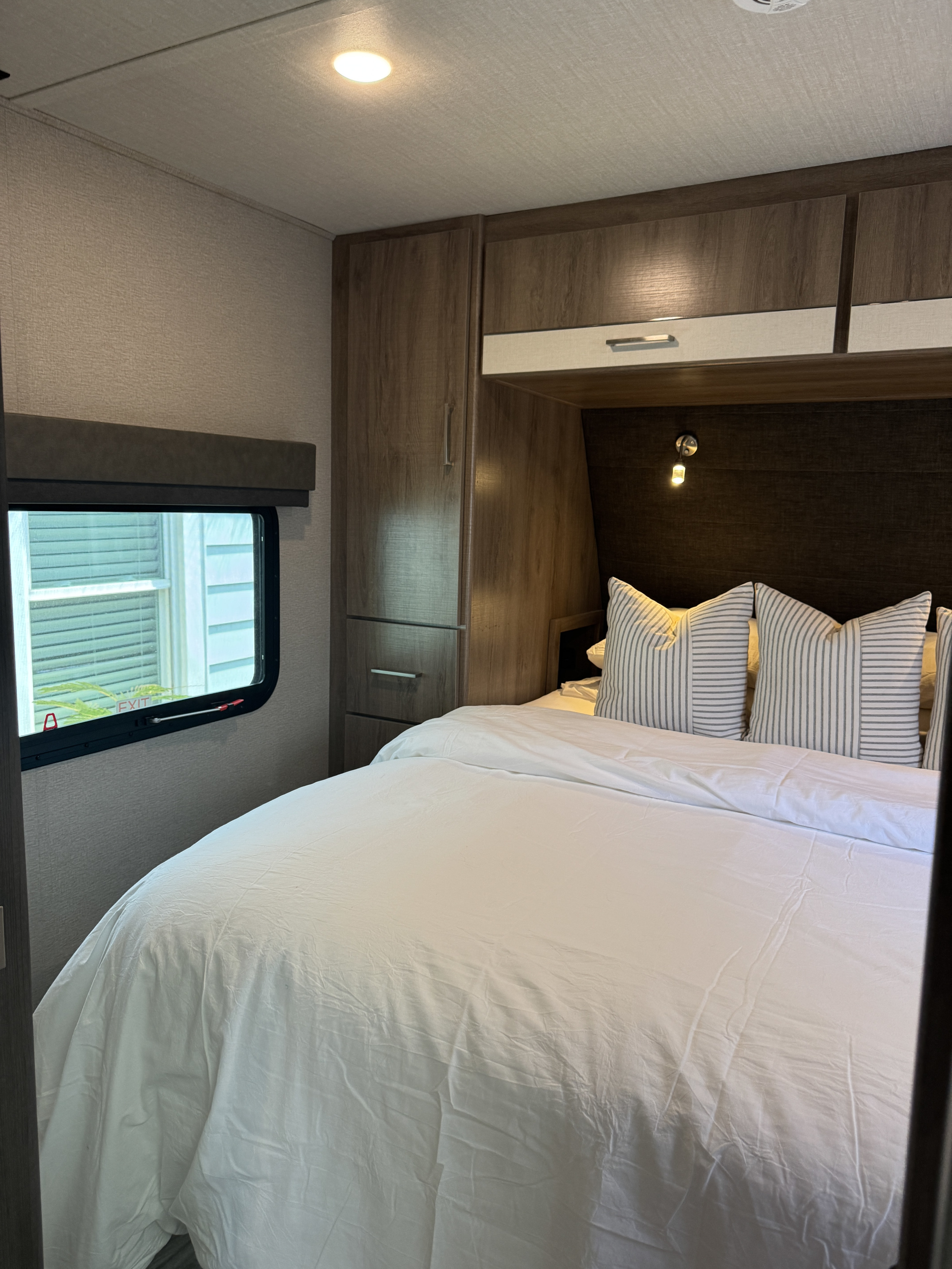 Inside a modern **rv rental** bedroom featuring a bed with white linen, striped pillows, wood cabinets, and a window.. Grand Design Imagine XLS 2023
