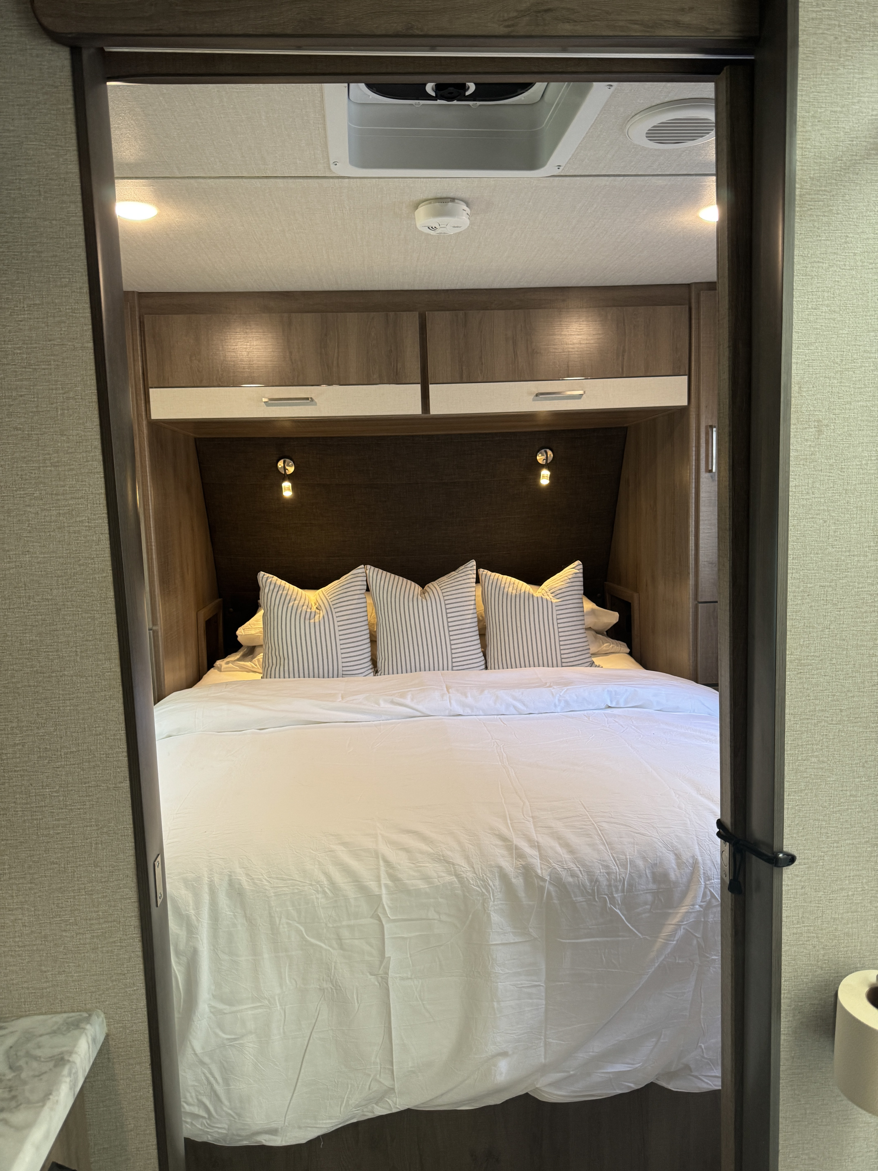 Interior view of an RV rental bedroom: cozy bed with striped pillows, overhead cabinets, and ceiling vent/smoke detector.. Grand Design Imagine XLS 2023