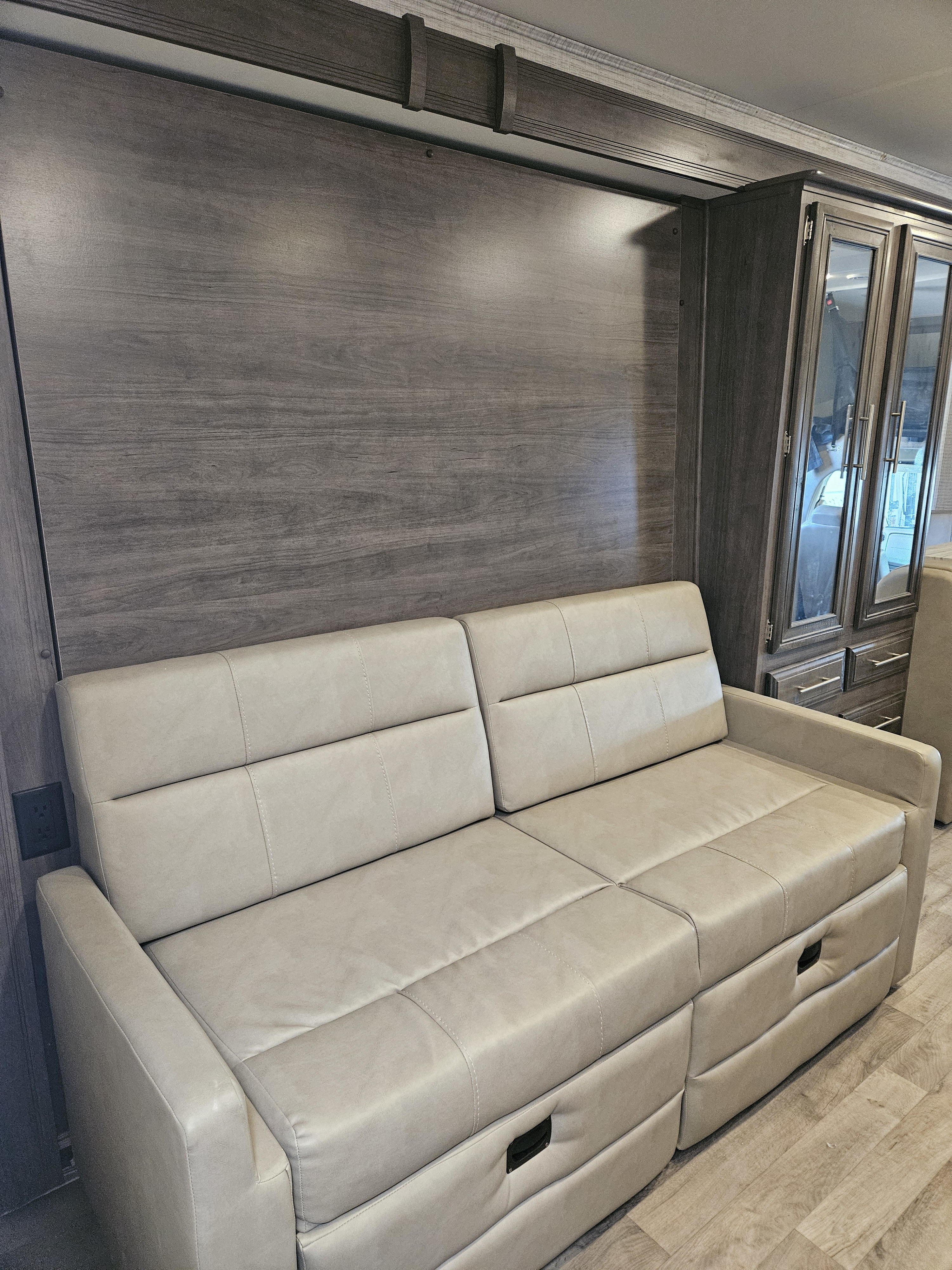 RV rental interior: a beige sofa against dark wood paneling, beside a matching storage cabinet with glass doors.. Jayco Redhawk 2024