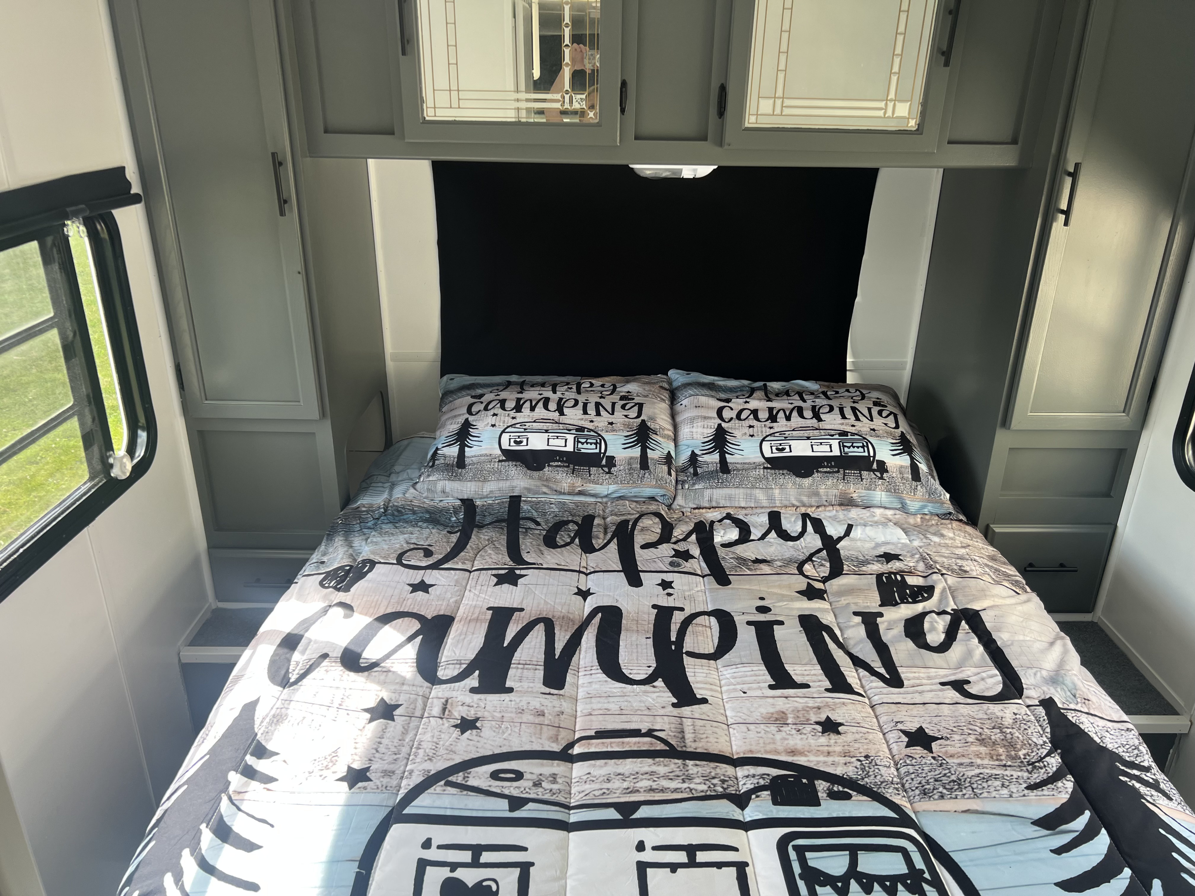 An RV rental's bedroom features a "Happy Camping" comforter, built-in side storage, and a window for scenic views.. Shasta Sprite 2002