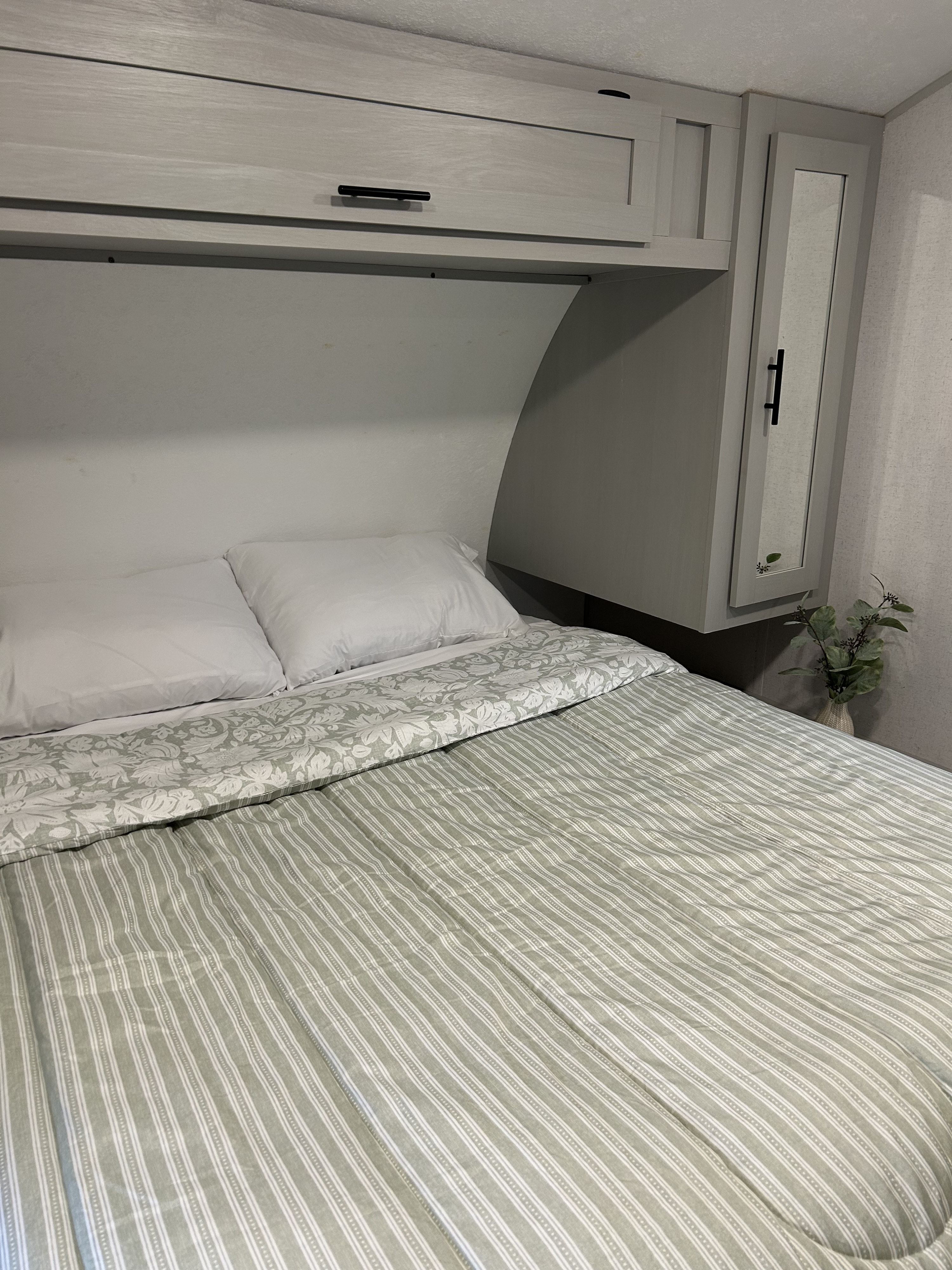Cozy RV rental bedroom features a striped comforter, white pillows, overhead storage cabinets, and a mirror.. Forest River Wildwood FSX 2021