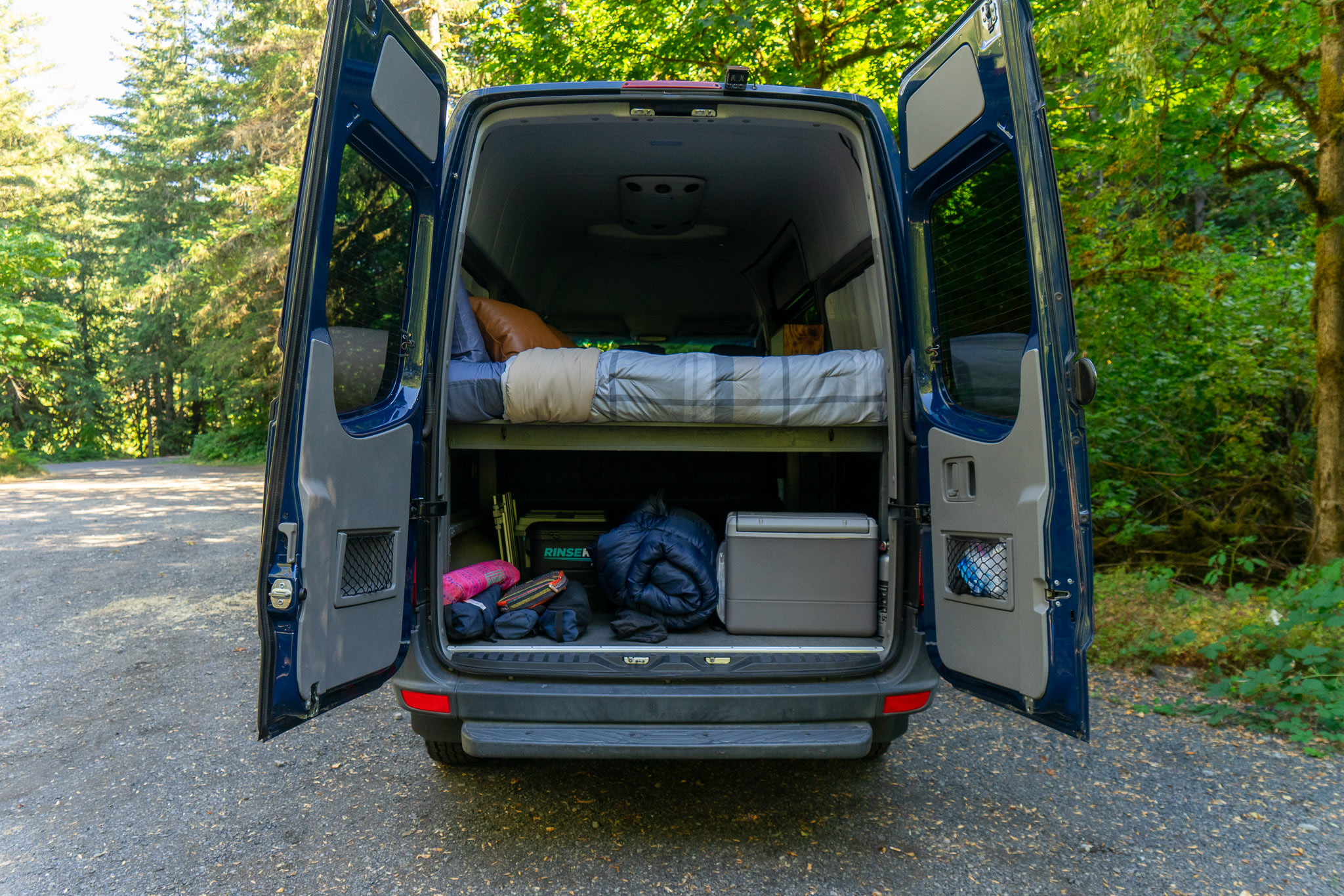 Open rear doors of a blue RV rental reveal a comfortable bed above and ample gear storage below for your next van life adventure.. Mercedes-Benz Sprinter 2016