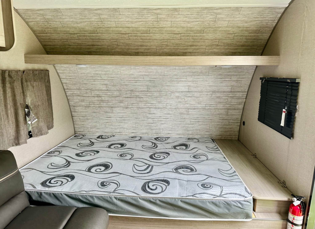 Cozy RV rental interior with a patterned mattress, wood-look walls, and compact sleeping area. Features a window with blinds.. Coachmen Catalina 2023