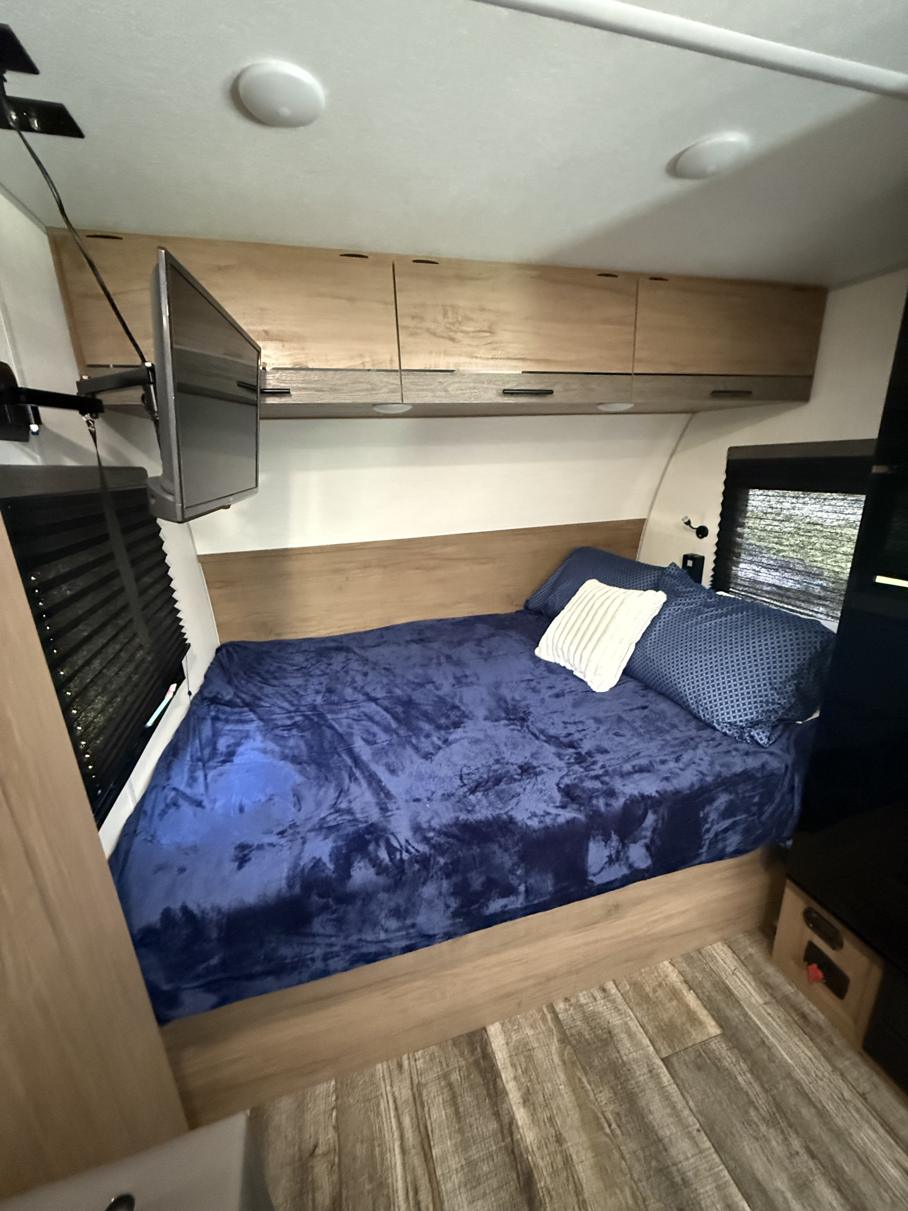 Inside an RV rental: a cozy bedroom with a blue blanket, wood-look floor, overhead storage, and a wall-mounted TV.. Forest River Campsite Reserve LS17 2025