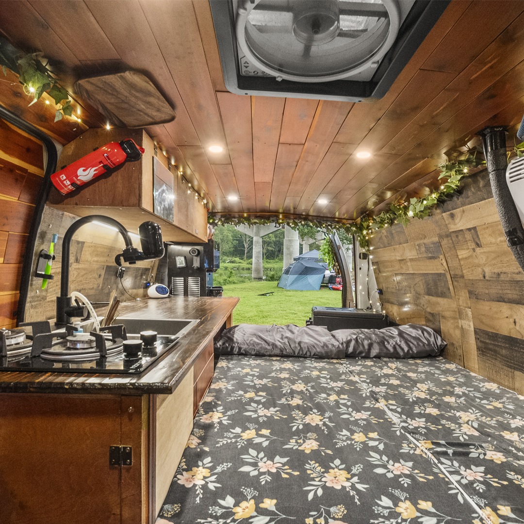 Cozy wood-paneled RV rental interior with kitchen, floral bed, and open back revealing a grassy campsite with tents.. Ford Econoline 150 2007