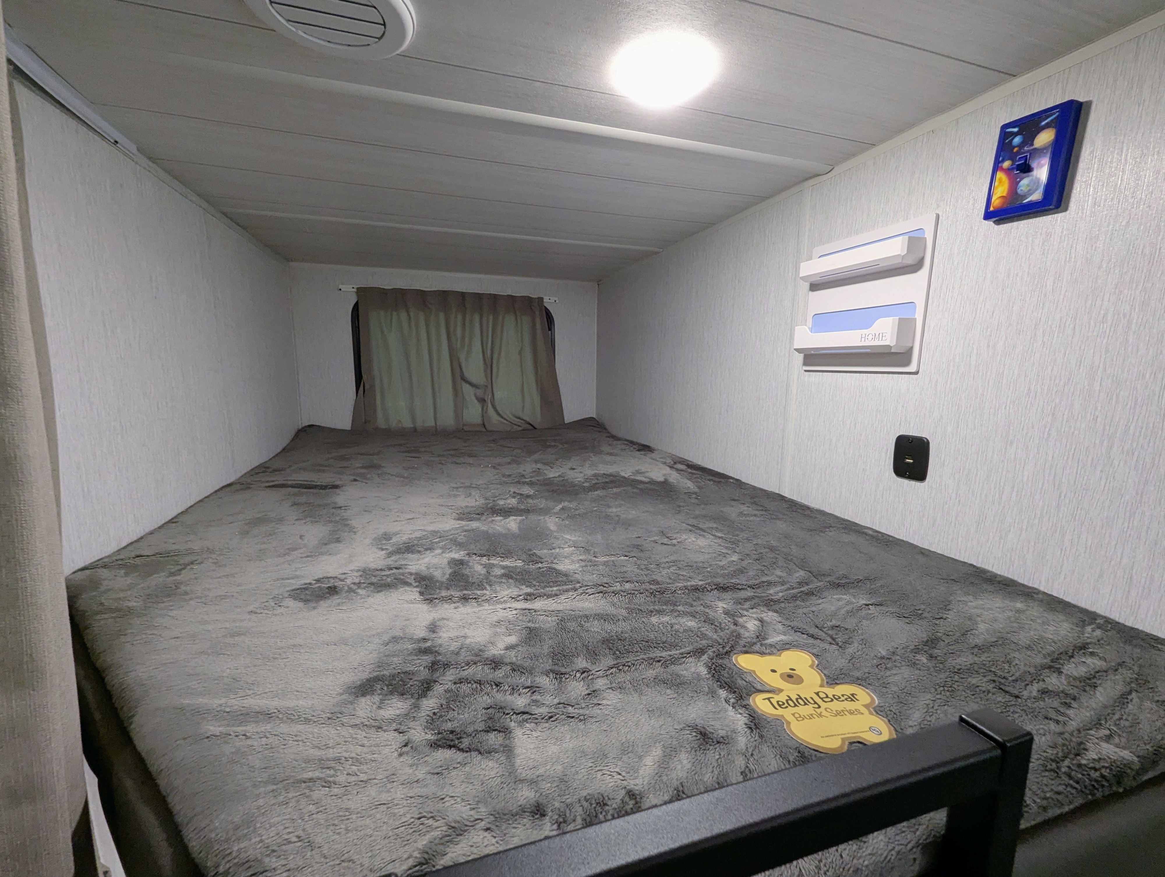 Inside a compact RV rental bunk, featuring a soft grey bed, window, light, and USB charger for comfortable travel.. Forest River Wildwood X-Lite 2024