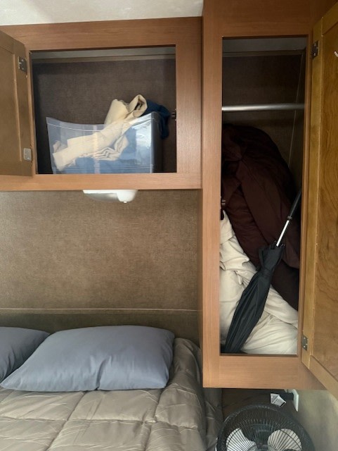 RV rental bedroom: Bed, two open overhead storage cabinets. One has a bin, the other holds clothes, umbrella. Fan on floor.. StarCraft Autumn Ridge 2013