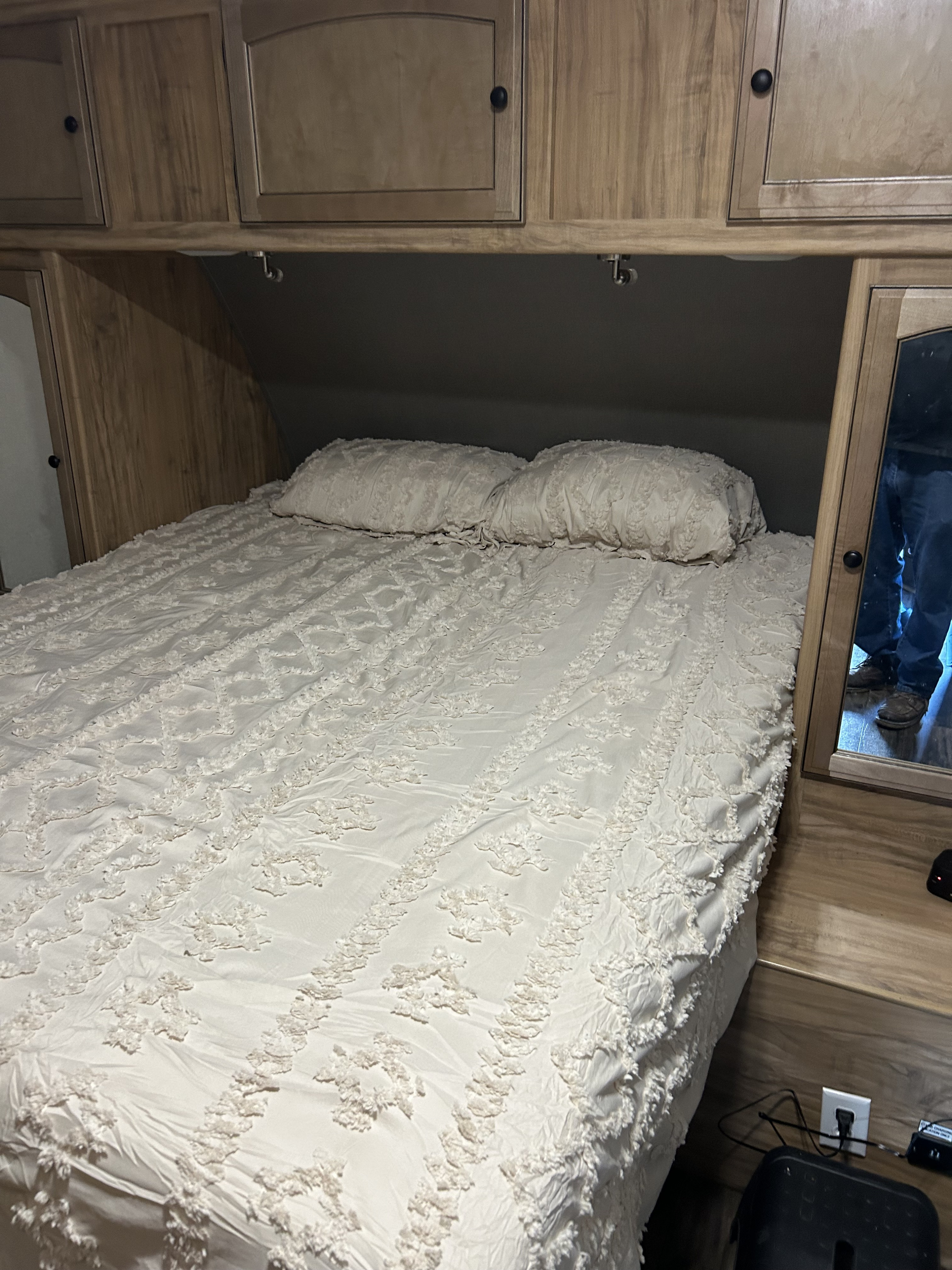 Cozy RV rental bedroom featuring a bed with textured white bedding, overhead wood cabinetry, and a full-length mirror.. Coachmen Freedom Express 2018