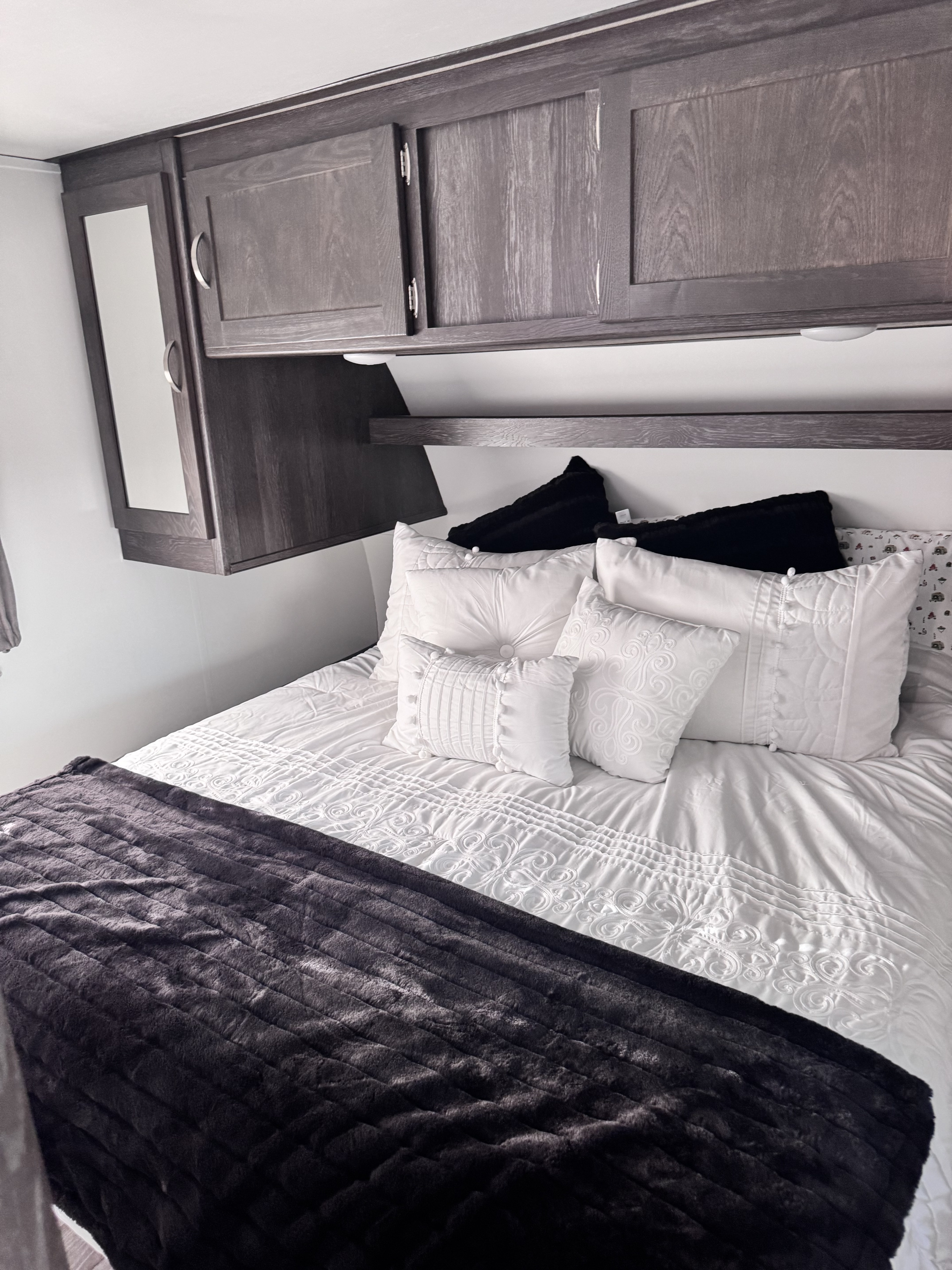 Enjoy a cozy RV rental bedroom featuring a white bed, decorative pillows, dark gray blanket, and overhead cabinets.. Keystone RV Outback Ultra-Lite 2018