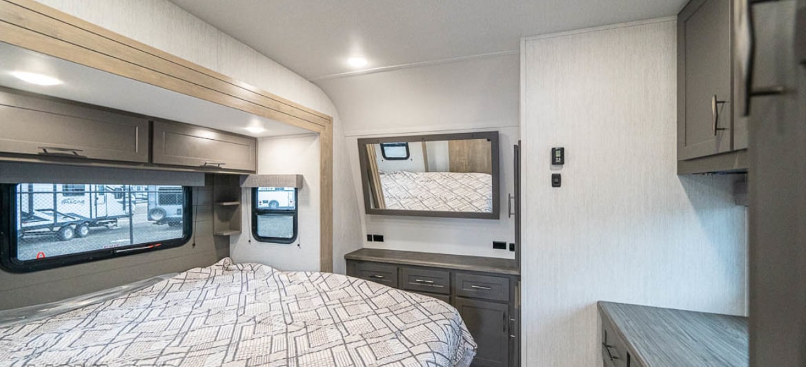Inside a modern RV rental, this bedroom features a comfortable bed, sleek overhead cabinets, and a dresser with a mirror.. Grand Design Momentum 2023