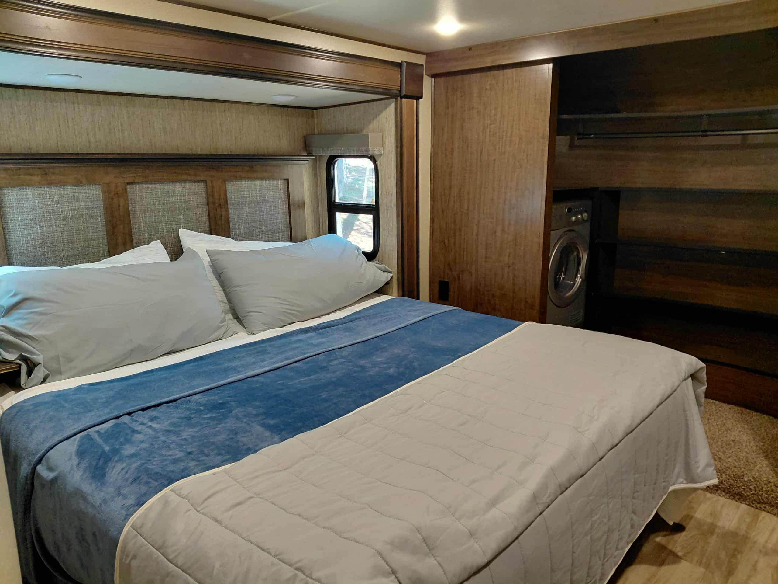 Inside an RV rental, a cozy bedroom with a bed, window, and closet revealing a convenient washer/dryer unit.. Grand Design Solitude 2019