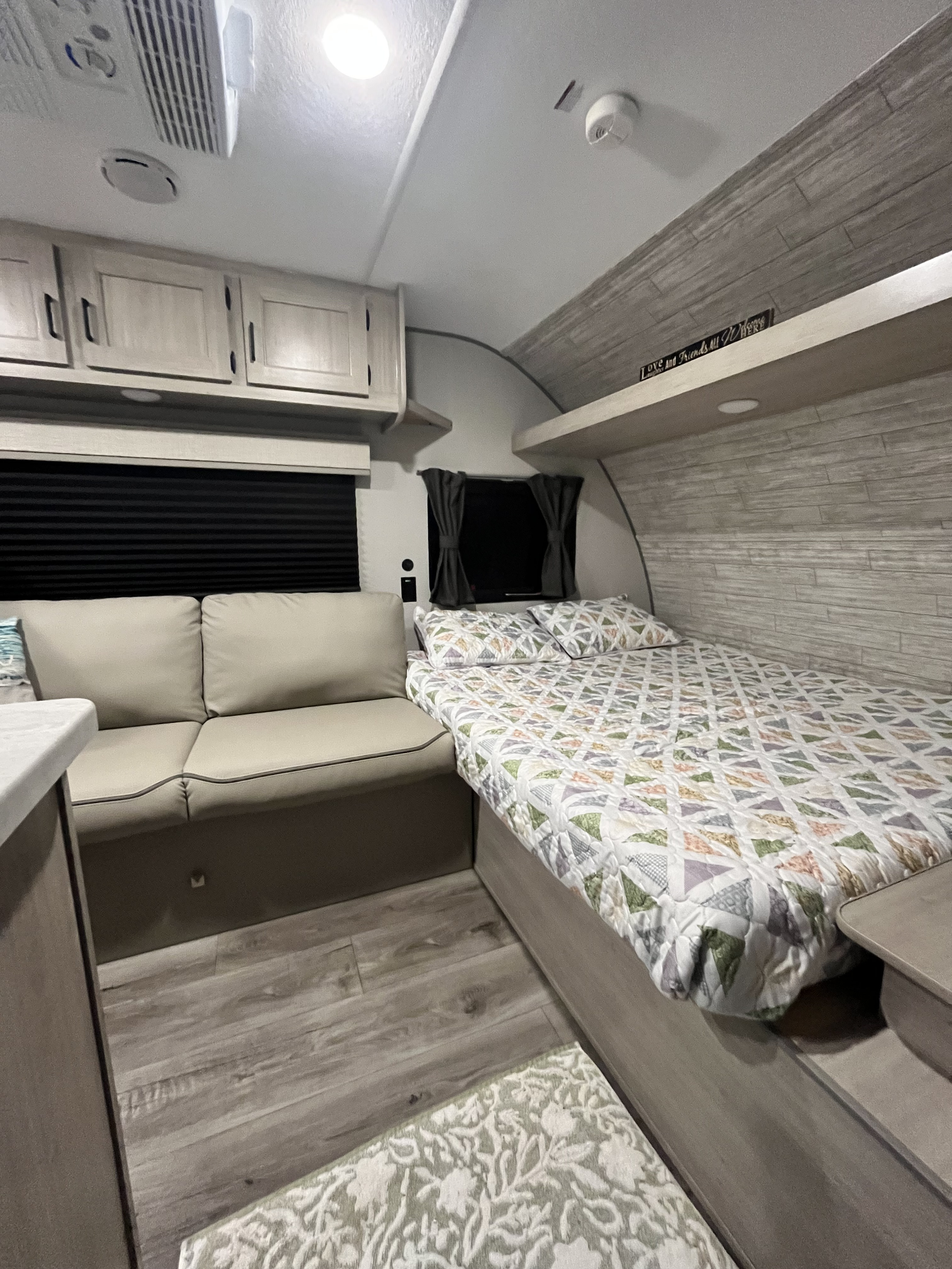 A compact RV rental interior features a patterned bed on the right, a cream sofa on the left, and light wood-tone cabinets.. Coachmen Catalina Summit 164BHX 2024