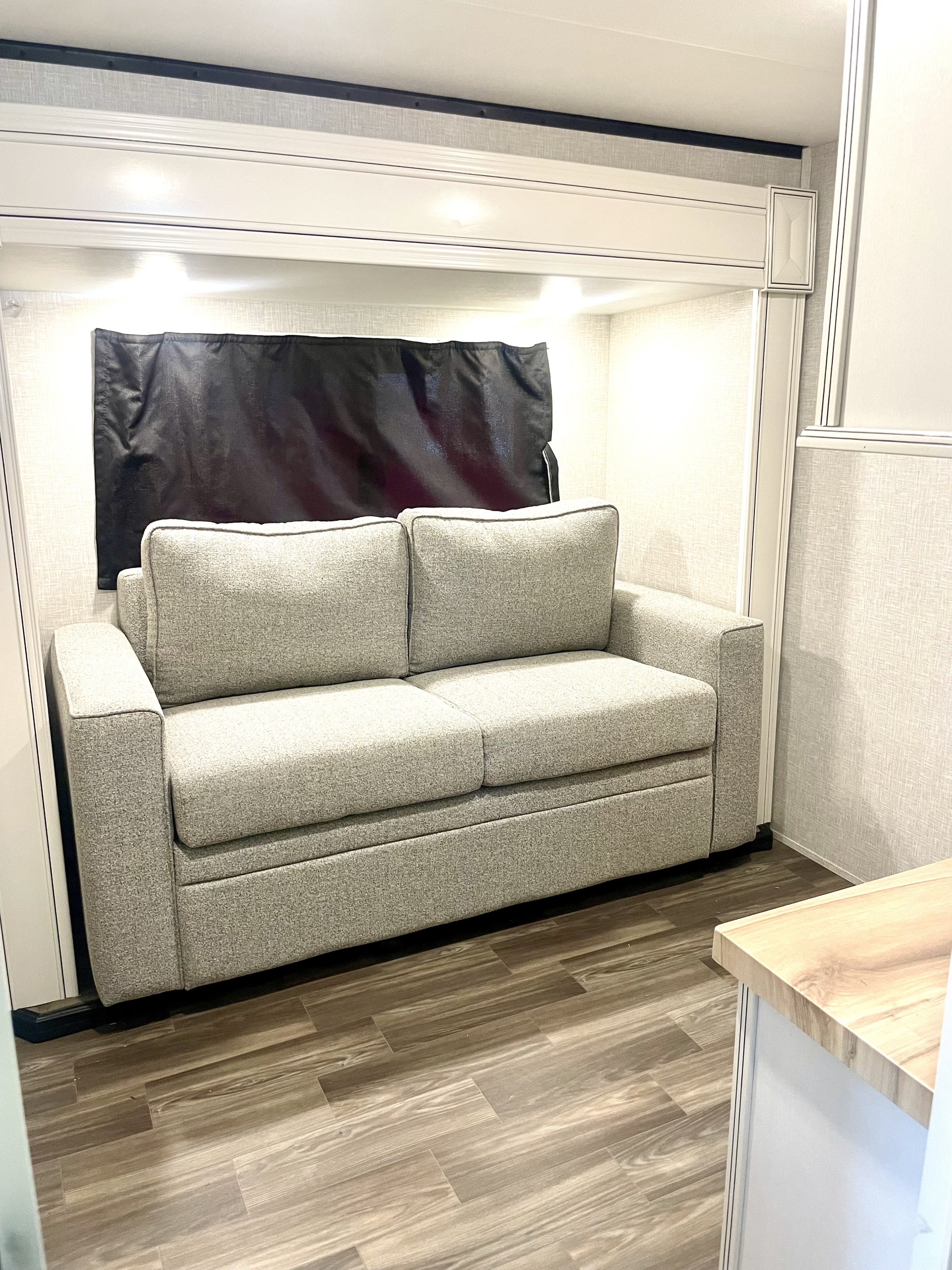 Inside an RV rental, a grey two-seater sofa with accent lighting and a blackout curtain. Wood-look flooring completes the cozy space.. Highland Ridge RV Open Range 2024
