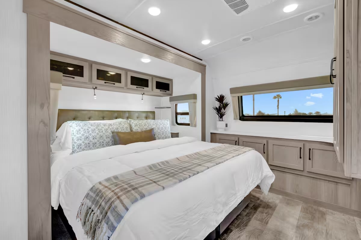 A bright, modern RV rental bedroom featuring a large bed with white linens, overhead storage, and windows showcasing a palm tree.. Forest River Flagstaff Classic 2024
