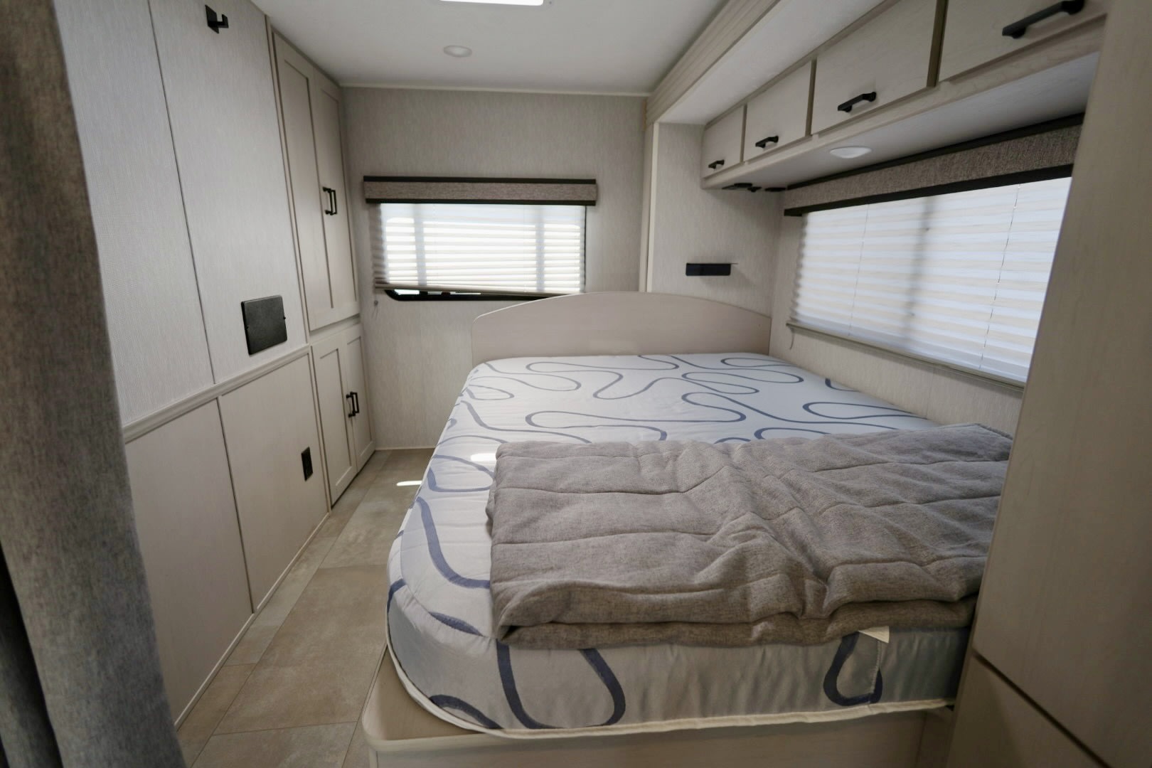 Cozy bedroom in an RV rental, featuring a comfortable bed, overhead cabinets, and windows with blinds for private travel.. Coachmen Freelander 2023
