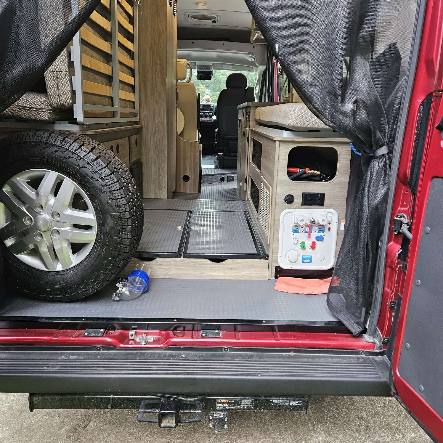 An RV rental camper van's interior, seen from the open side door. Features include a bed frame, spare tire, and water utility panel.. Winnebago Solis 59PX 2022