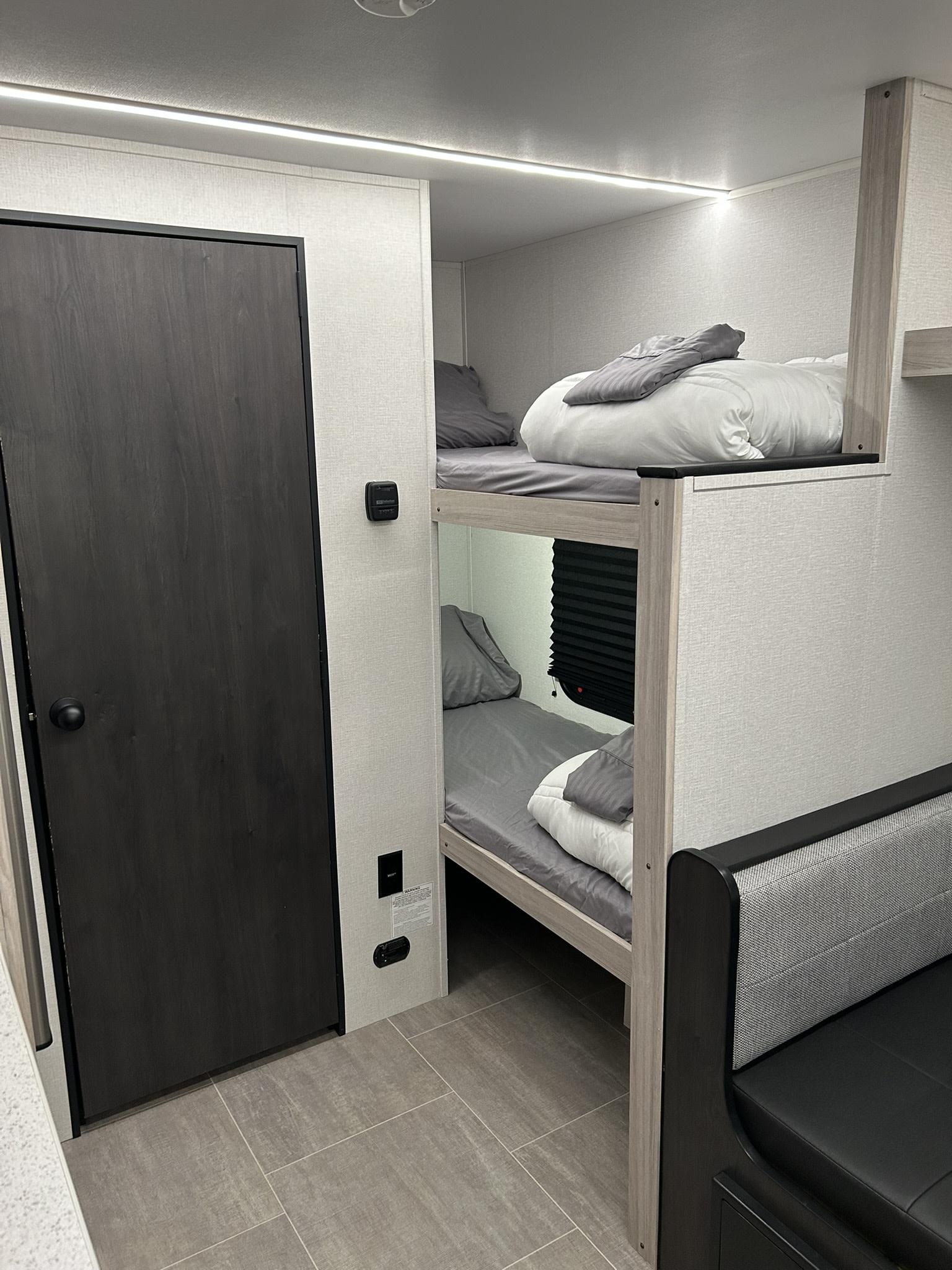 Modern RV rental interior: cozy bunk beds with grey bedding, a dark wood door, LED lighting, and compact living space.. Heartland Prowler 2025