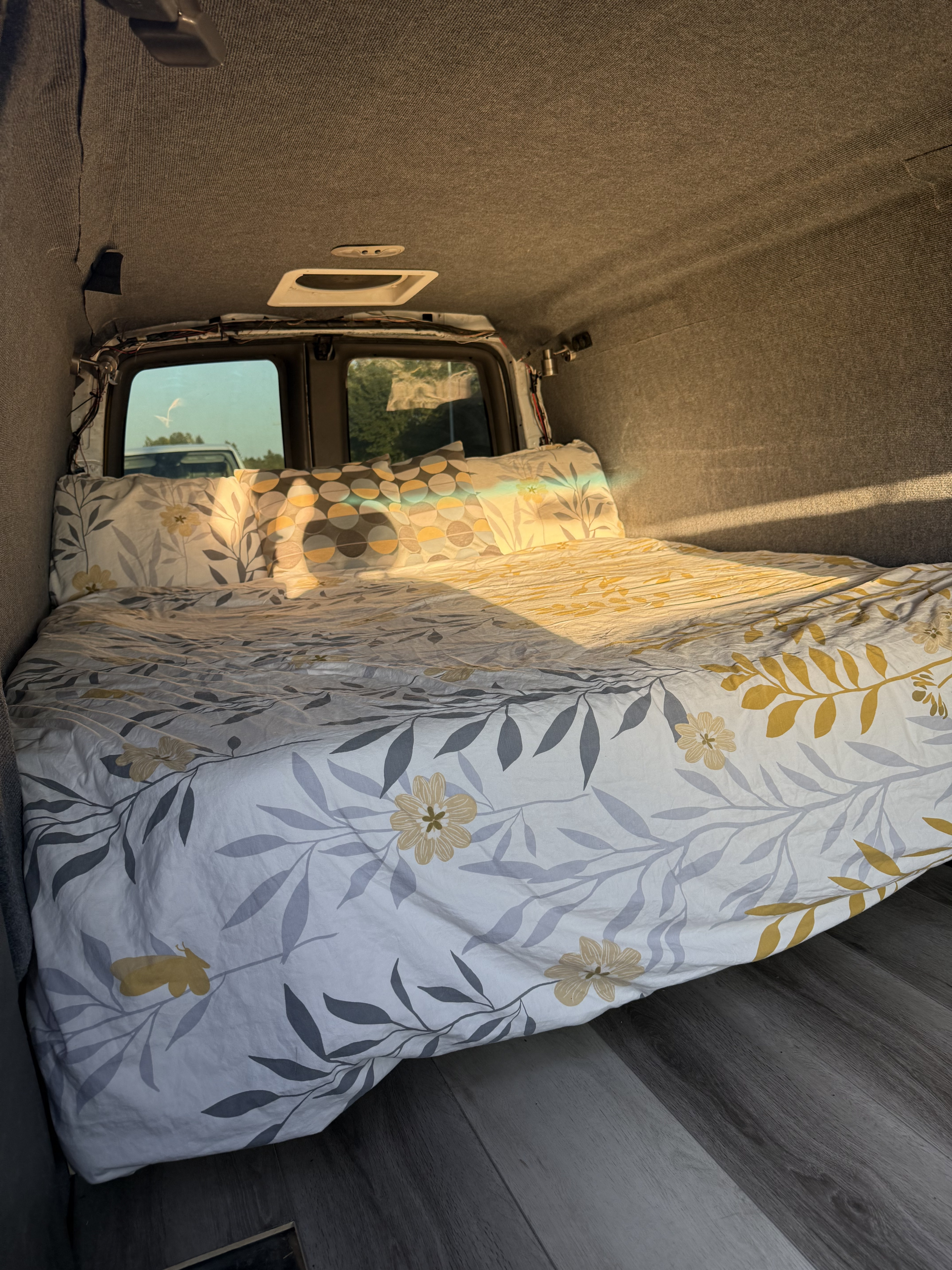 Comfortable RV rental bed with bright floral bedding inside a van, illuminated by sunlight through rear windows.. Gmc Savanna 2006