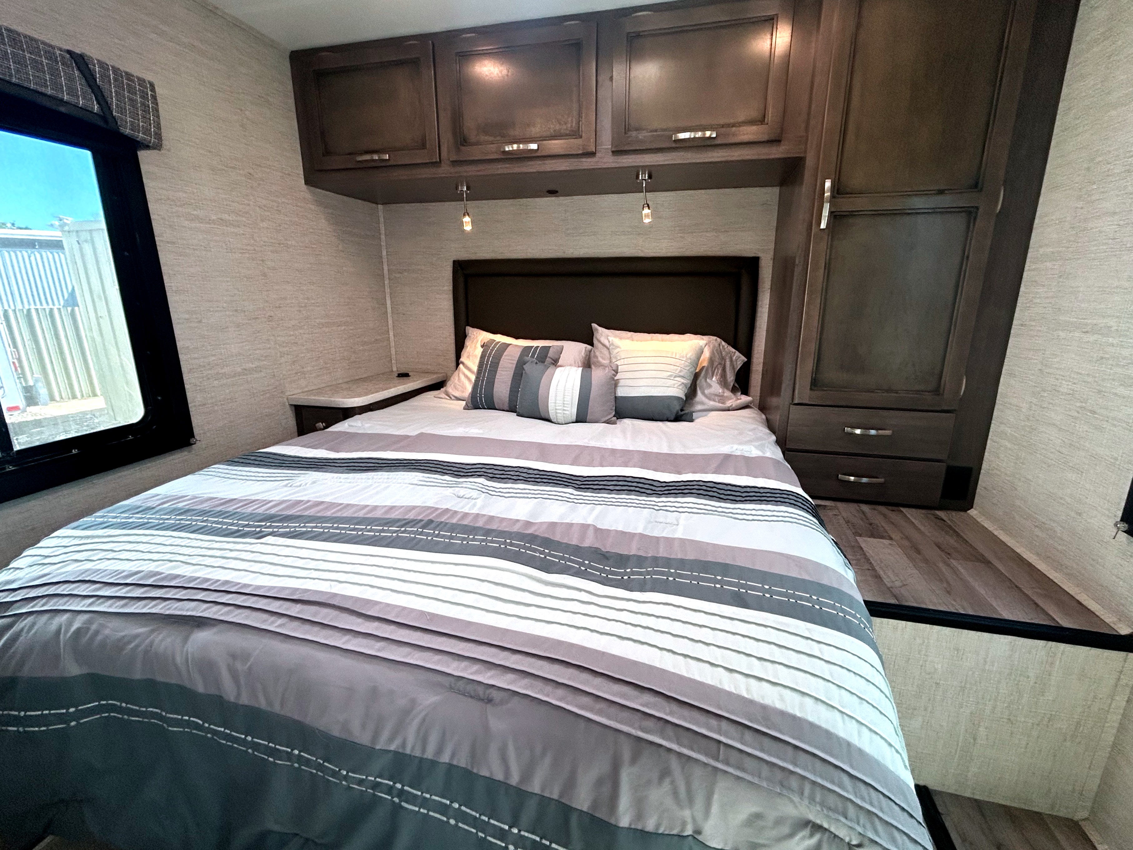 A modern RV rental bedroom featuring a comfortable bed with striped bedding, dark wood cabinets, and a window.. Jayco Redhawk SE 2023