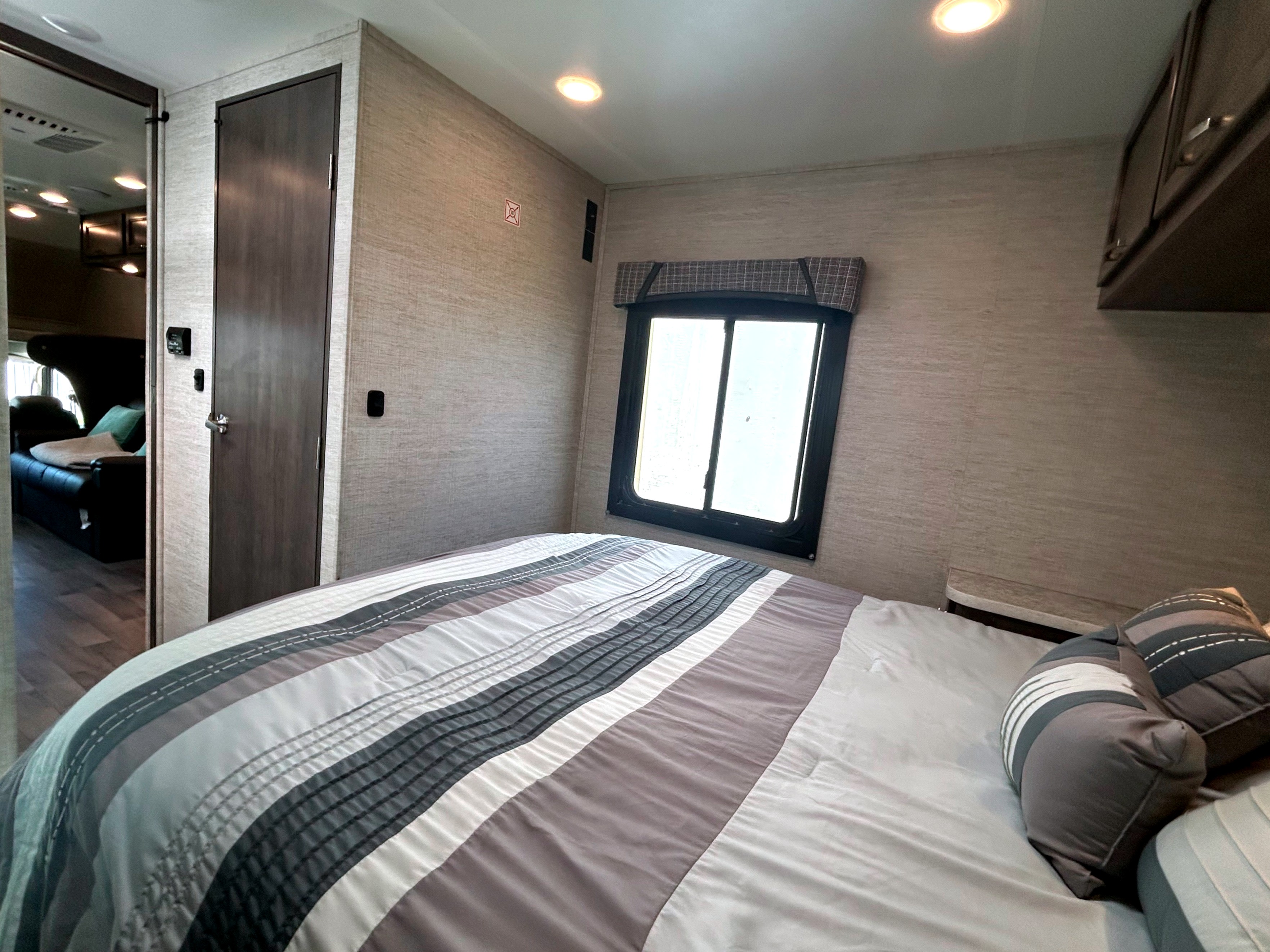 Cozy RV rental bedroom featuring a comfortable bed with striped comforter, window, and adjacent door. Ideal for your travels.. Jayco Redhawk SE 2023
