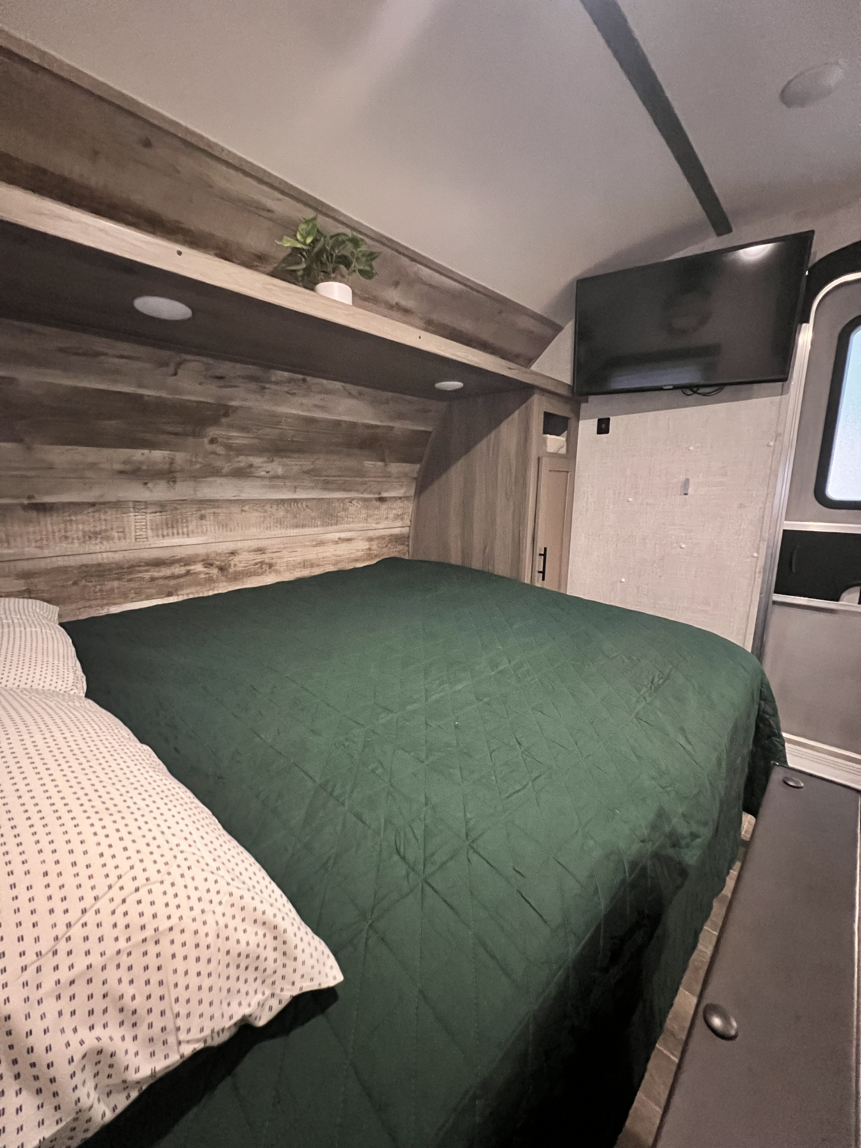 RV rental bedroom: dark green quilted bed, wood-paneled walls, shelf with a plant, and a wall-mounted TV. Cozy space.. Gulf Stream Ameri-Lite Super Lite 2021