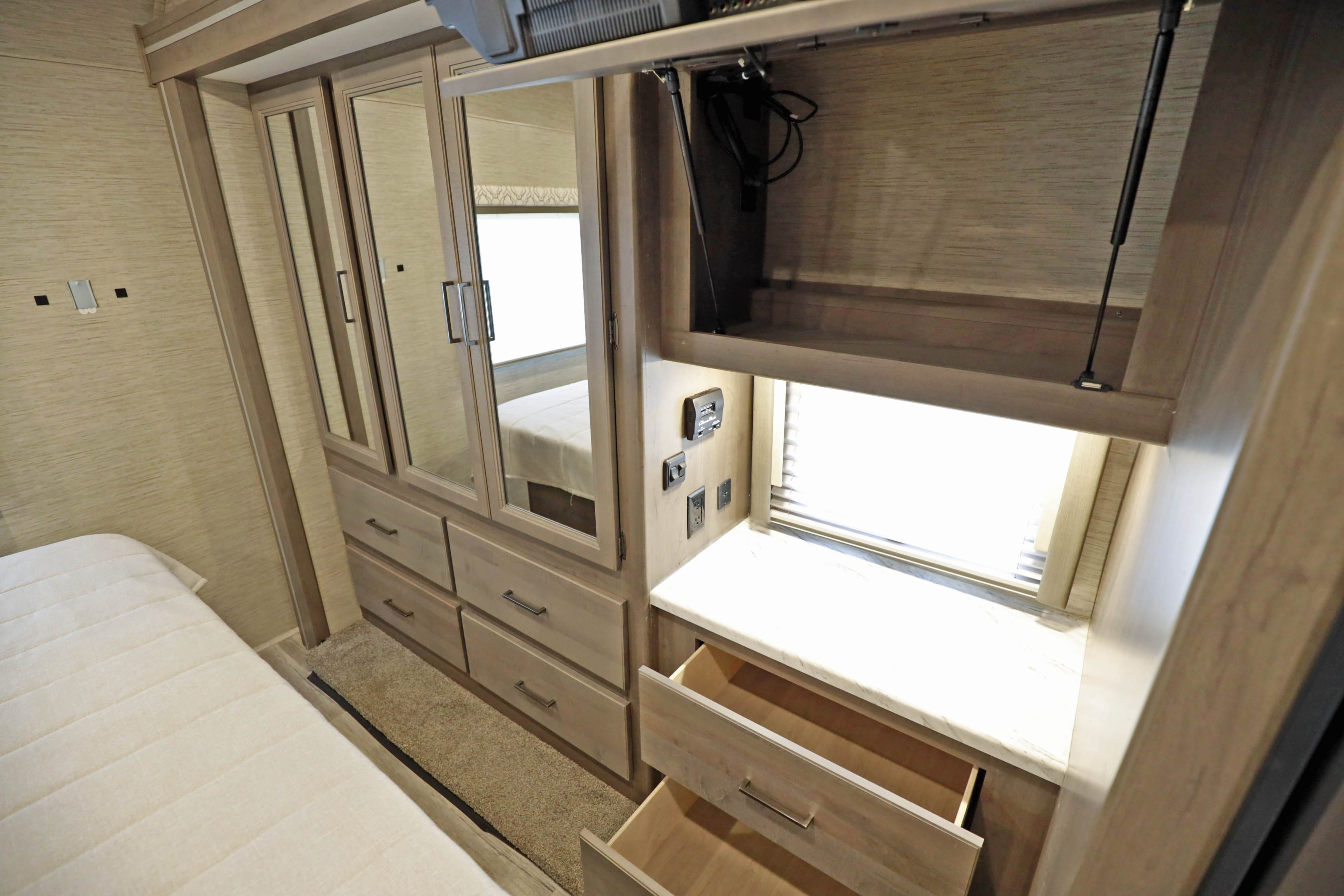 A light-toned RV rental bedroom features a bed, mirrored wardrobe, numerous drawers, and a built-in storage unit with a countertop and open drawers.. Classe A 31 pieds avec extensions 2021
