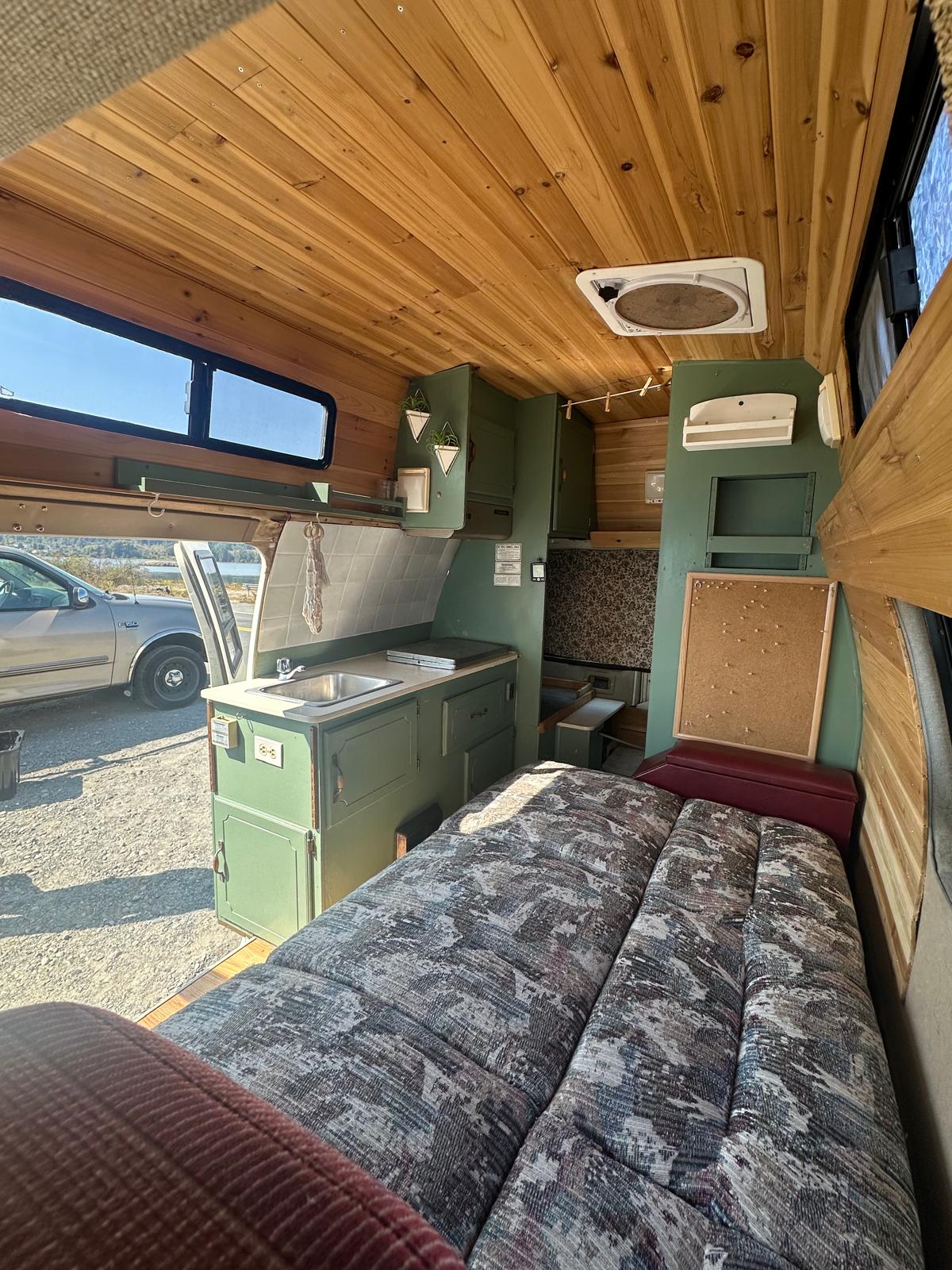 Inviting RV rental interior: wood-paneled ceiling, comfy bed, green kitchenette, and bright window view outside.. Ford Econoline 150 1989