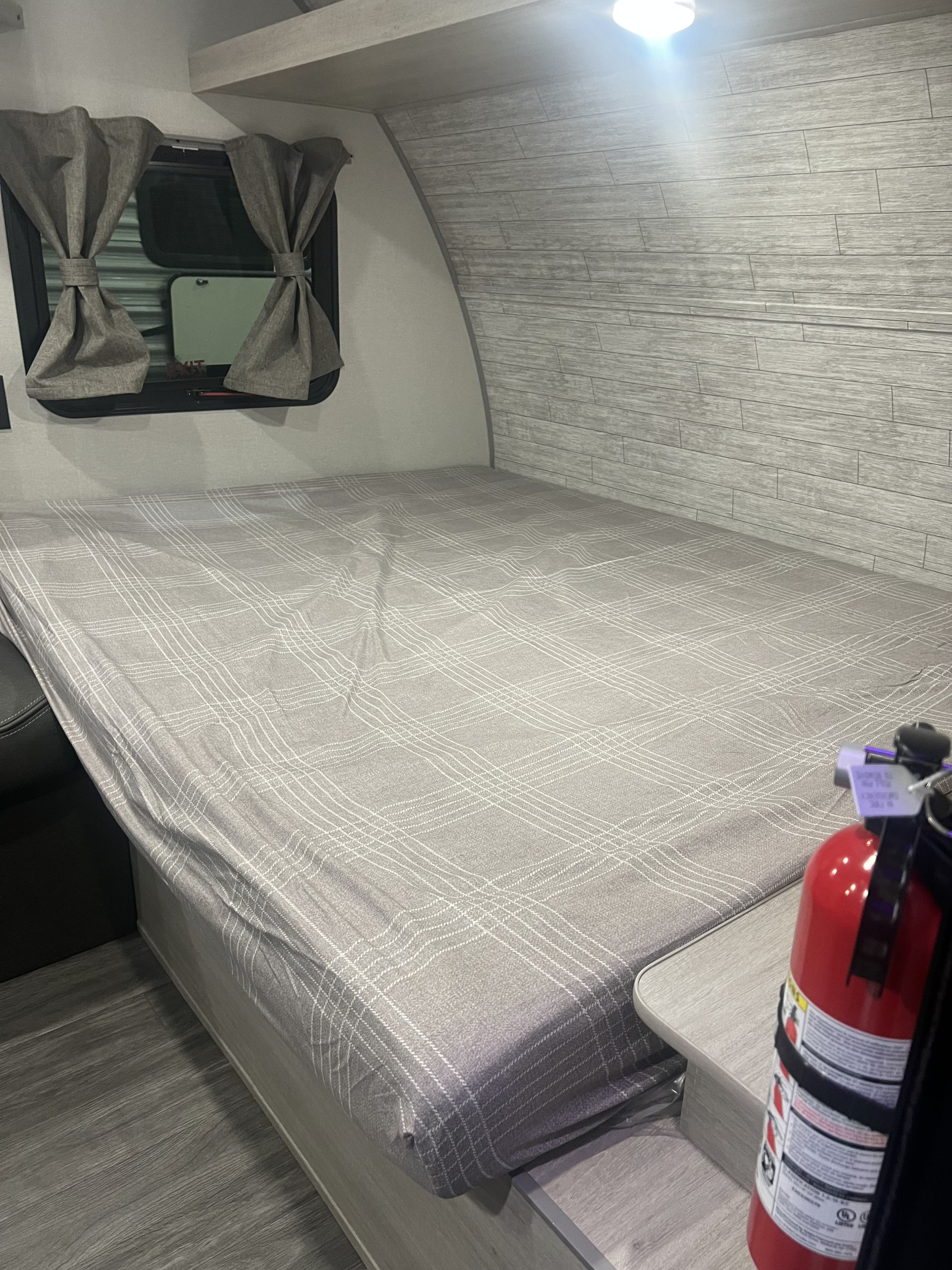 Inside an RV rental, a cozy bedroom features a bed with plaid sheet, window curtains, and a safety fire extinguisher.. Forest River Aurora 2025