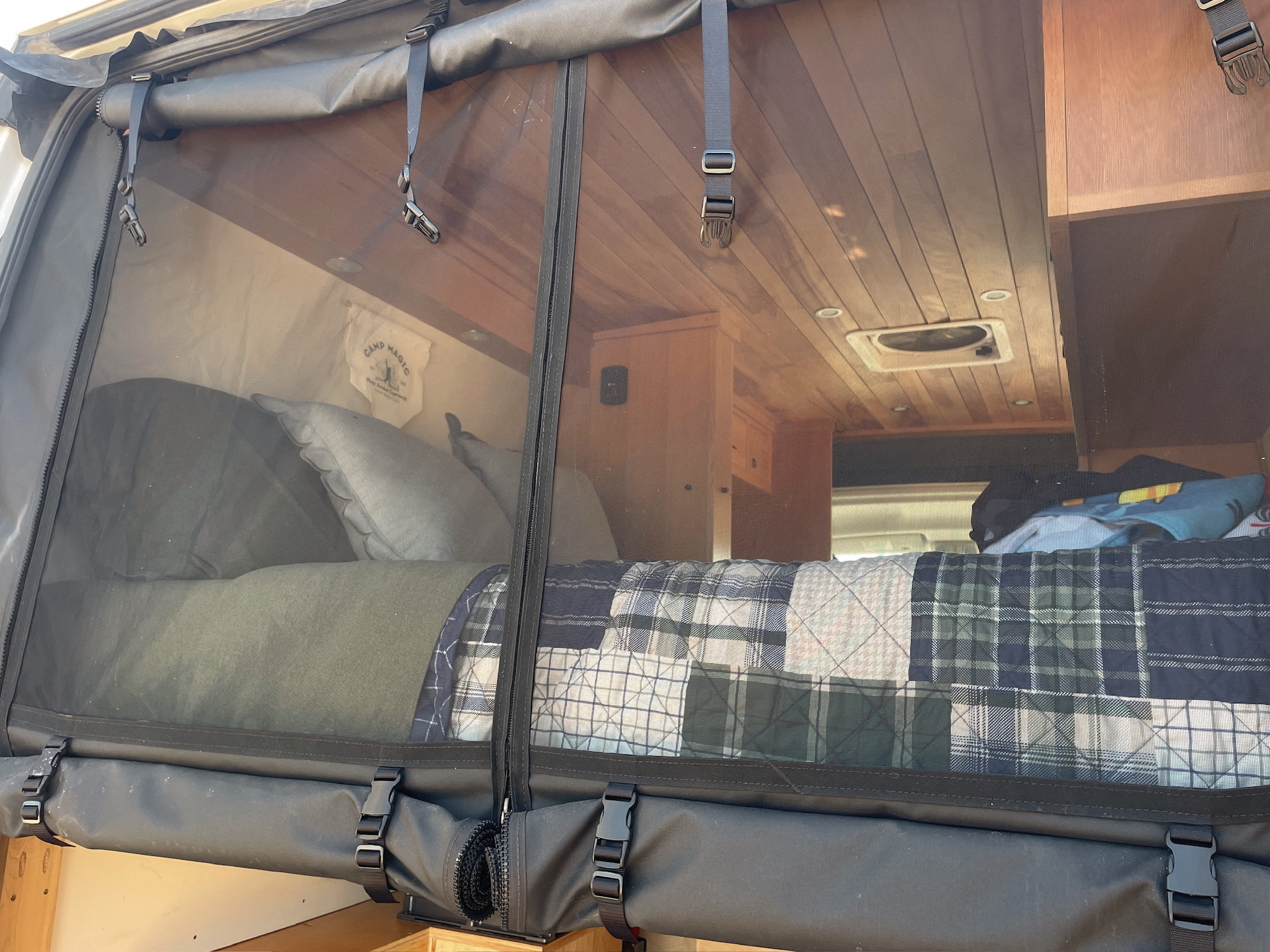 View into a cozy RV rental bedroom. A bed with plaid quilt, wood ceiling, & fan are visible through a protective mesh.. Dodge 1500 2018