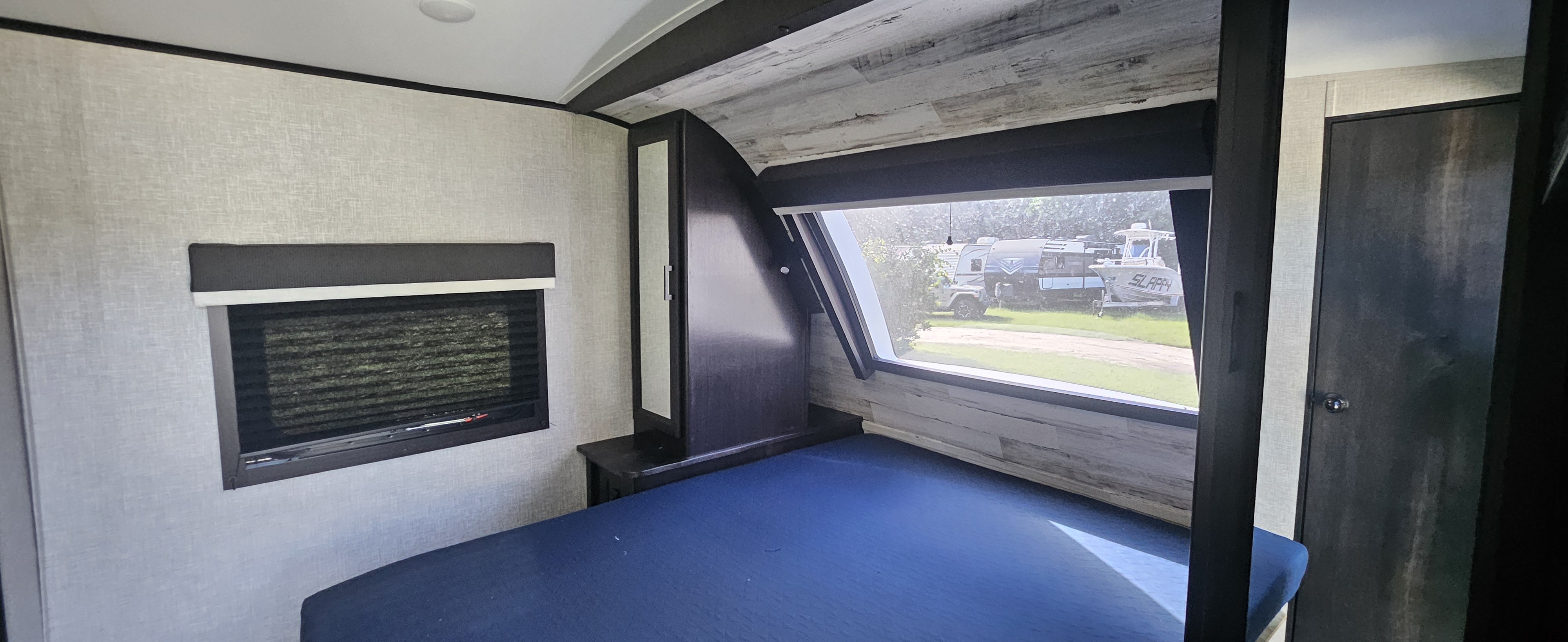 Cozy RV rental bedroom with a blue bed, side window with blinds, and front window showing an RV and 'SLAPPY' boat outside.. Jayco White Hawk 2022