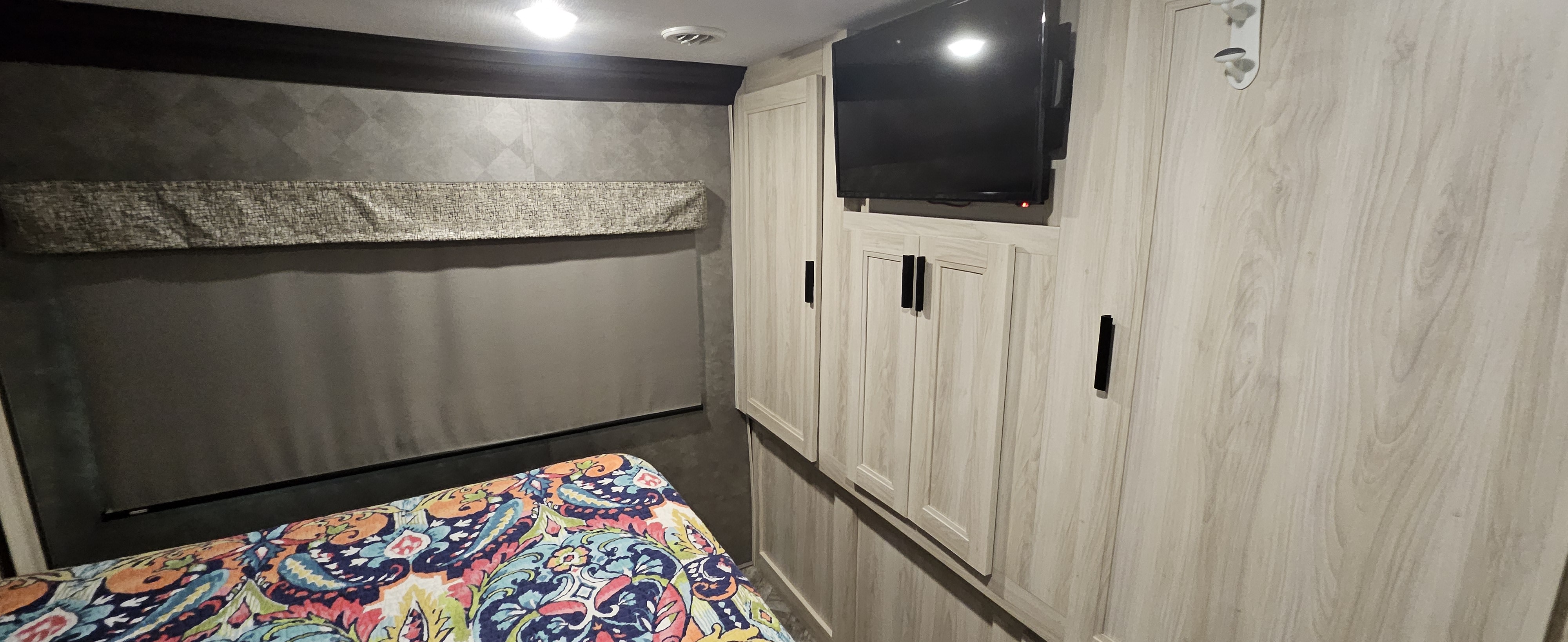 Inside an RV rental bedroom: vibrant bedding, sleek light wood cabinetry with black handles, a pull-down shade, and mounted TV.. Forester Classic 2021