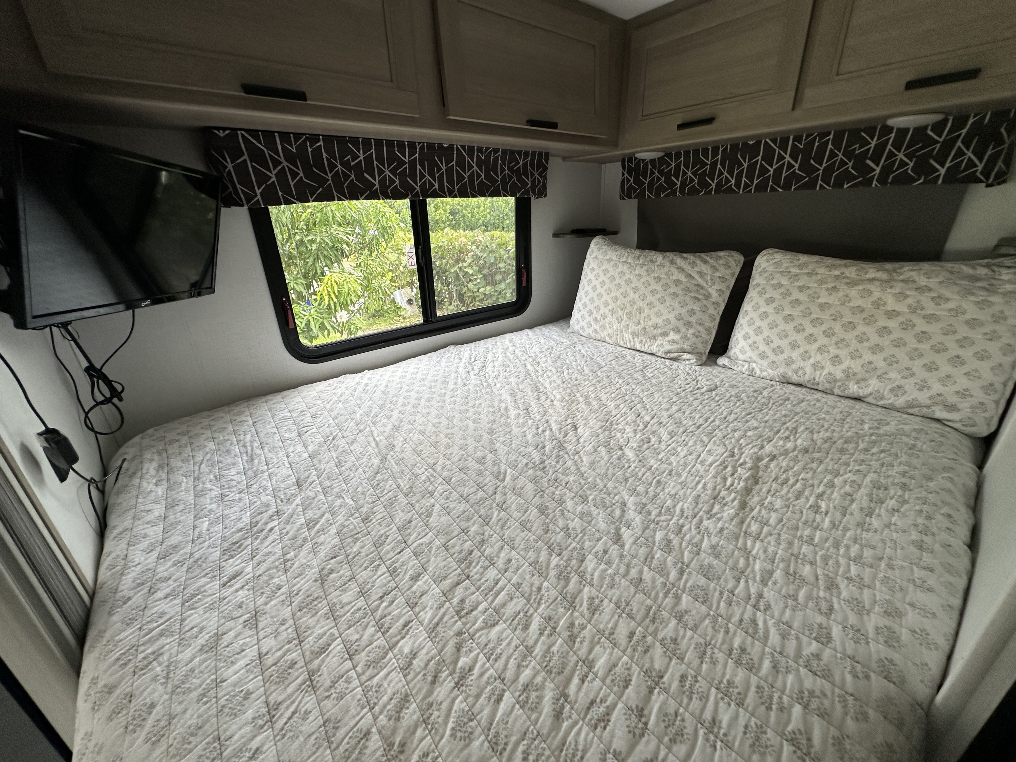 Cozy RV rental bedroom with patterned white bedding, a window overlooking trees, a mounted TV, and overhead cabinets.. Forest River Sunseeker LE 2024