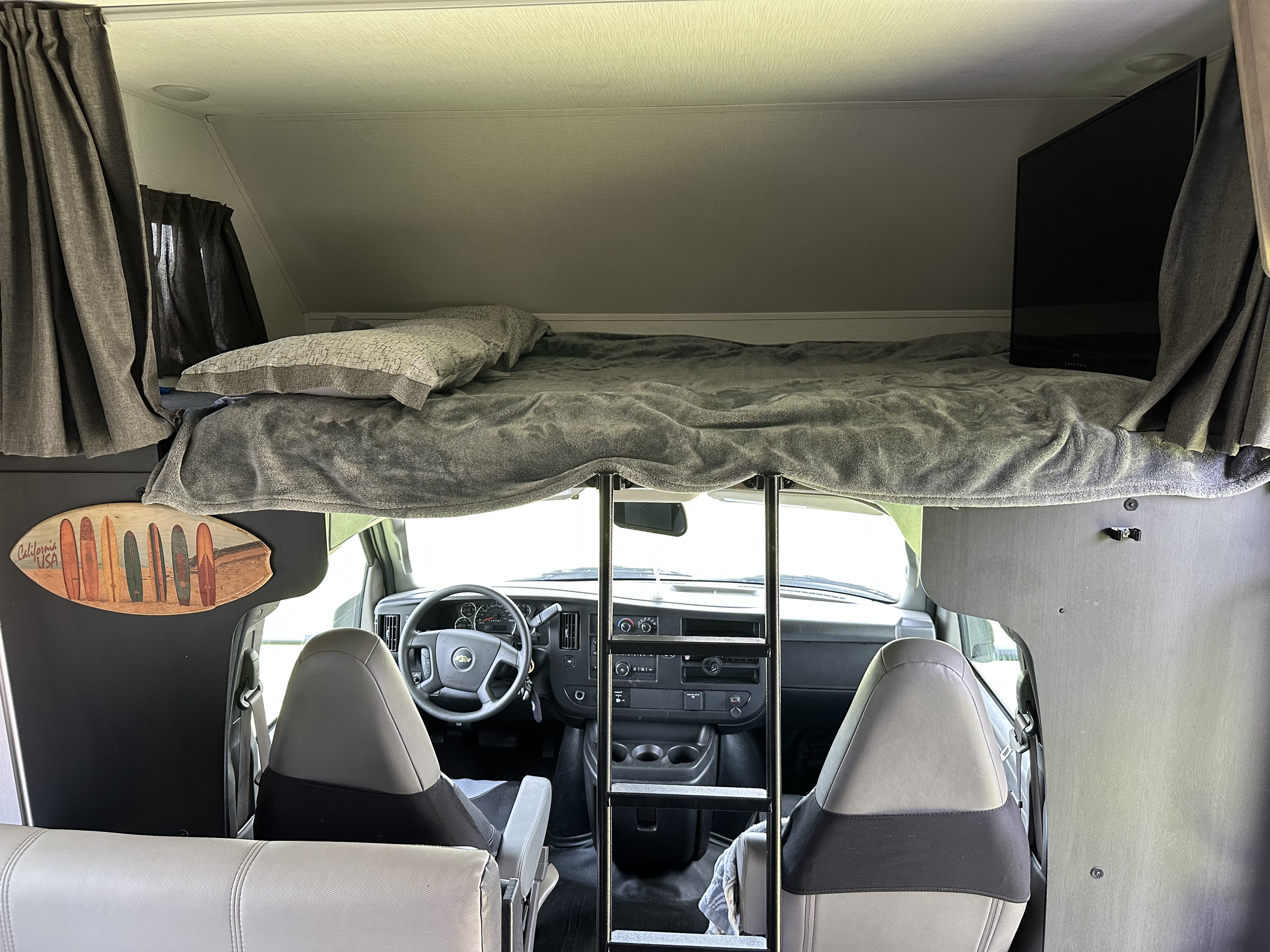 An RV rental interior showing a cozy over-cab bed with a TV, ladder, and the front driver's area.. Forest River Sunseeker LE 2024