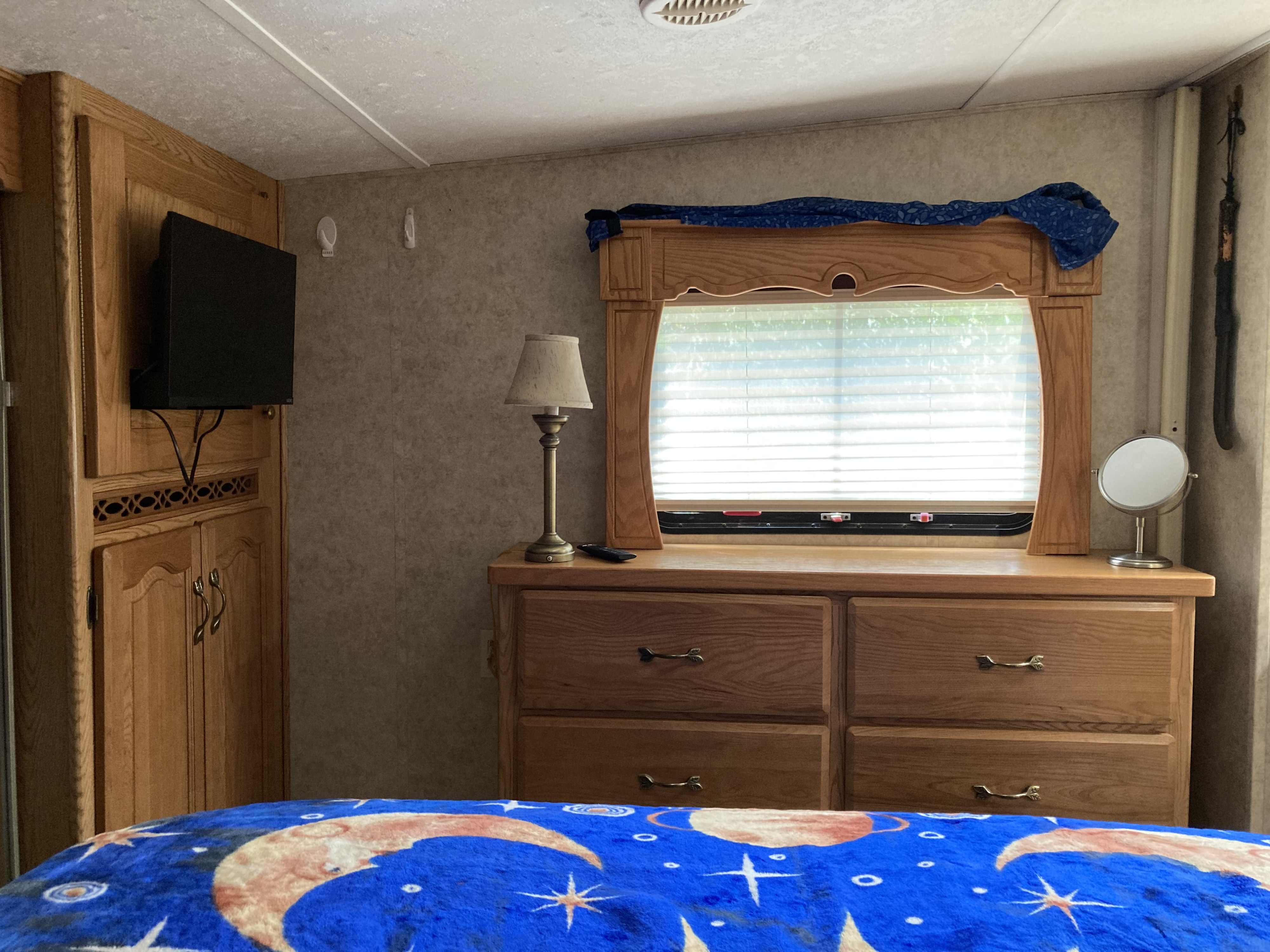 Inside a cozy RV rental bedroom. Features include wooden cabinets, a TV, a dresser with a window, and a bed with a blue, celestial blanket.. Keystone 2955RL 2006