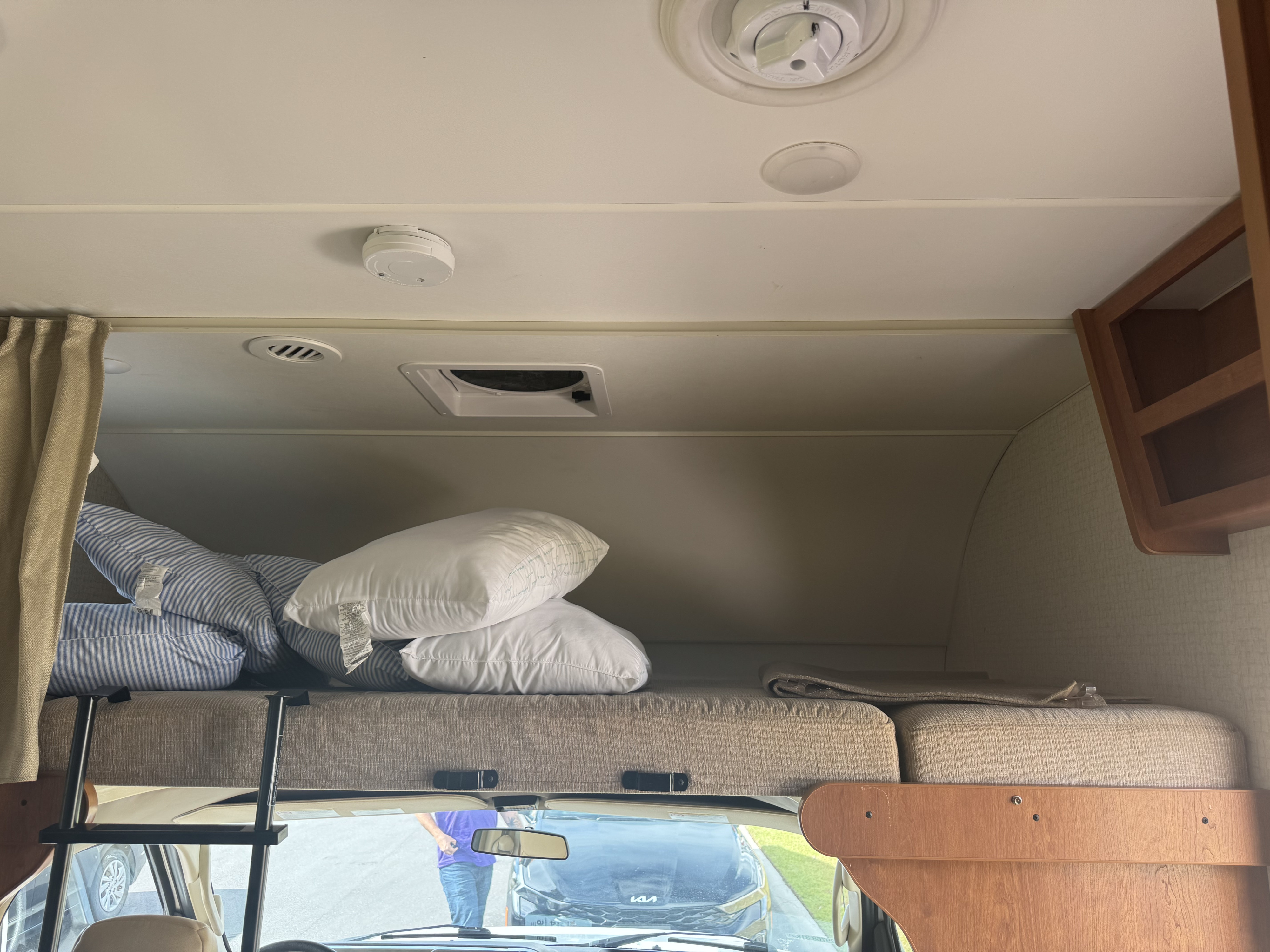 Cozy cab-over bunk in an RV rental, featuring pillows, a ladder, ceiling vents, and a smoke alarm in the overhead area.. Winnebago Other 2016