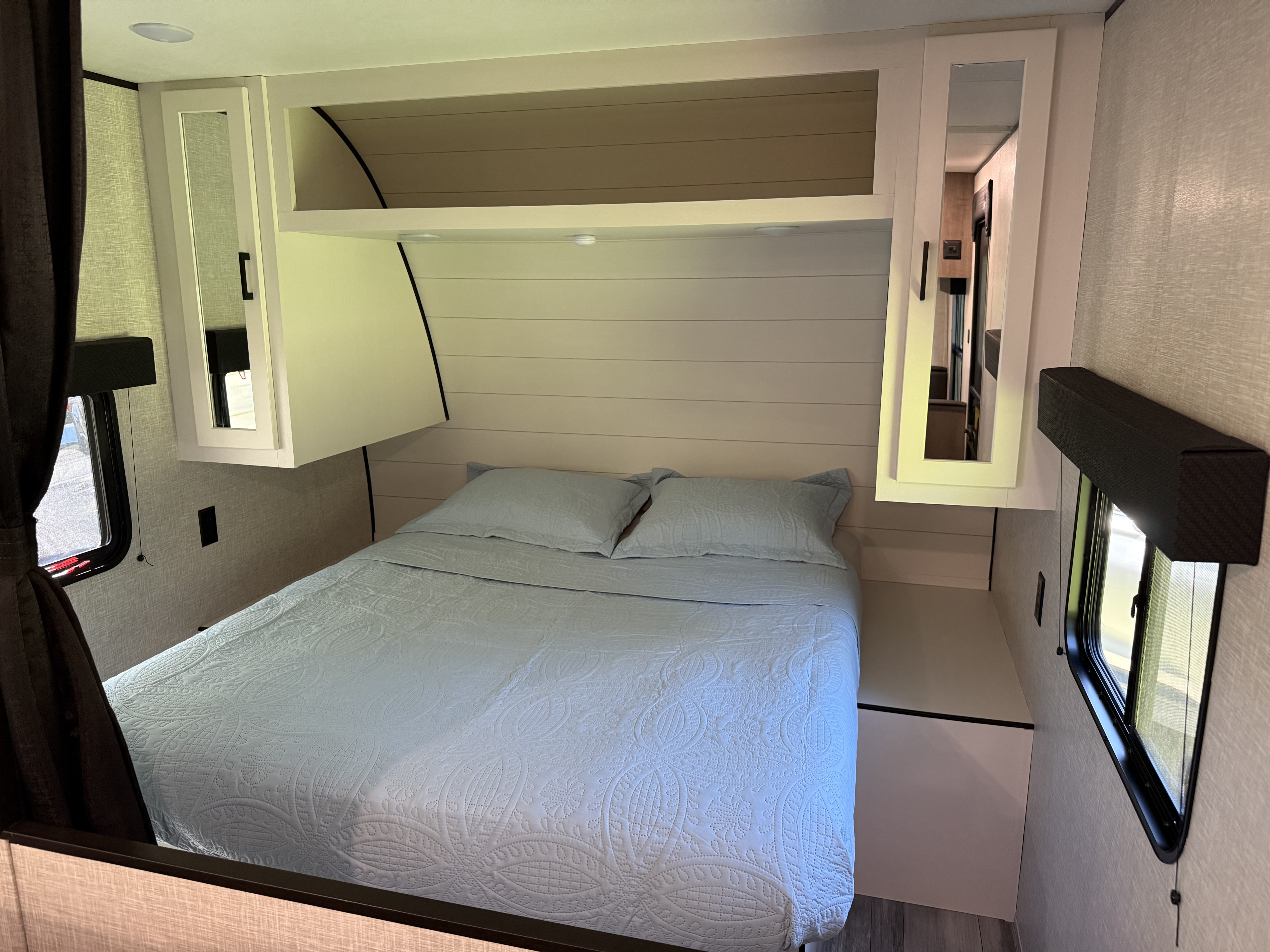 A clean, compact RV rental bedroom with a blue quilted bed, mirrored cabinets, and side windows for natural light.. Jayco Jay Flight SLX 2024