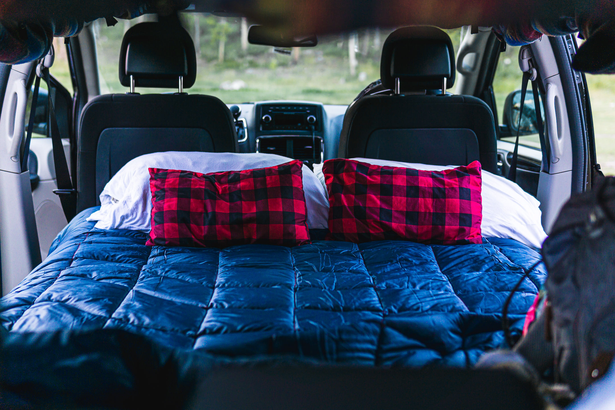 Ready for a road trip! This cozy RV rental's interior boasts a comfortable bed with a blue comforter and red plaid pillows.. Dodge Converted Dodge Caravan 2020