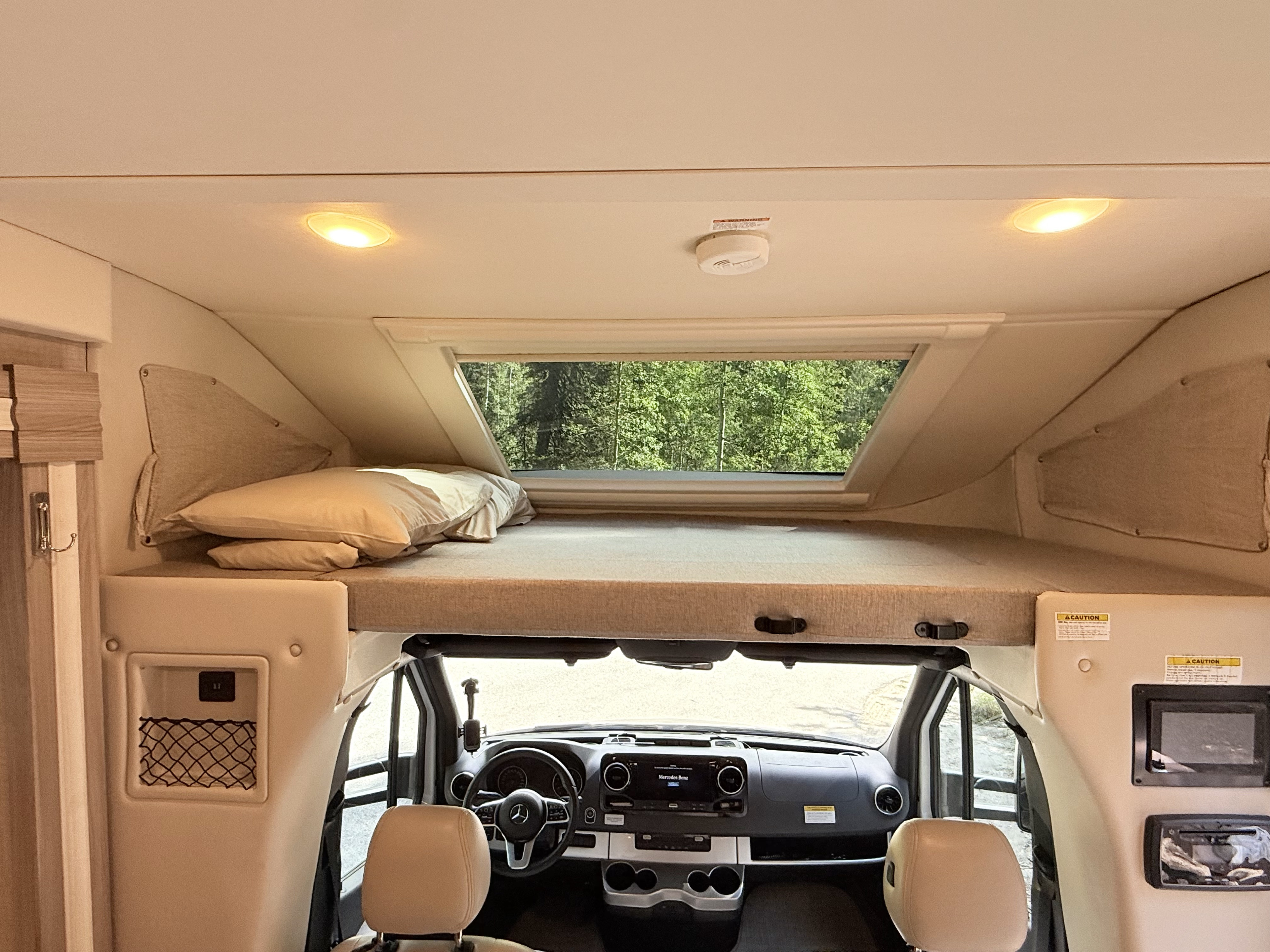 Inside an RV rental, showing the overhead bunk bed, cockpit, and window overlooking a forest. Perfect for road trips.. Thor Delano 2022