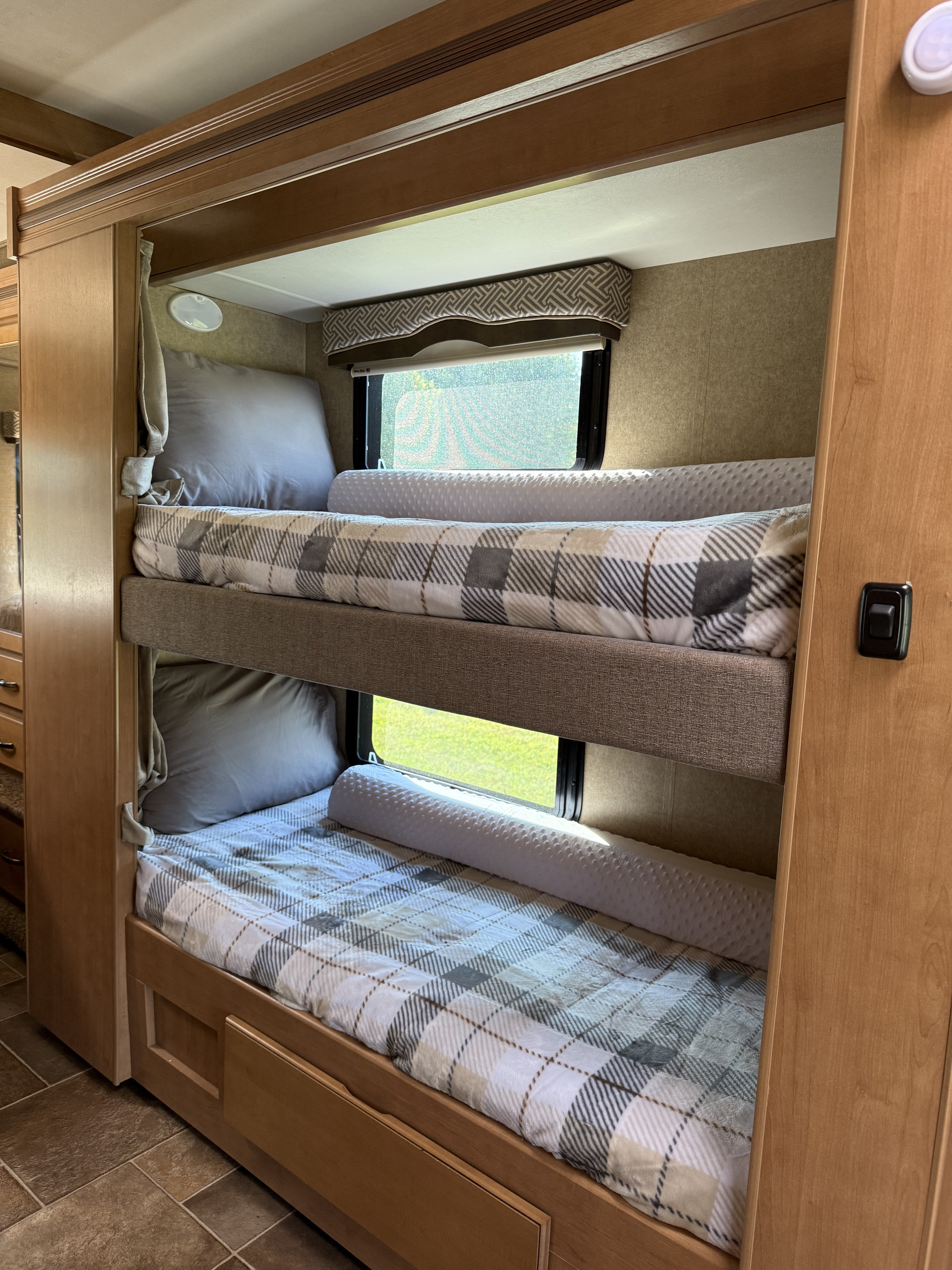 Cozy RV rental bunk beds with plaid blankets, pillows, and windows showing outdoor views, perfect for family camping.. Thor Chateau 2017