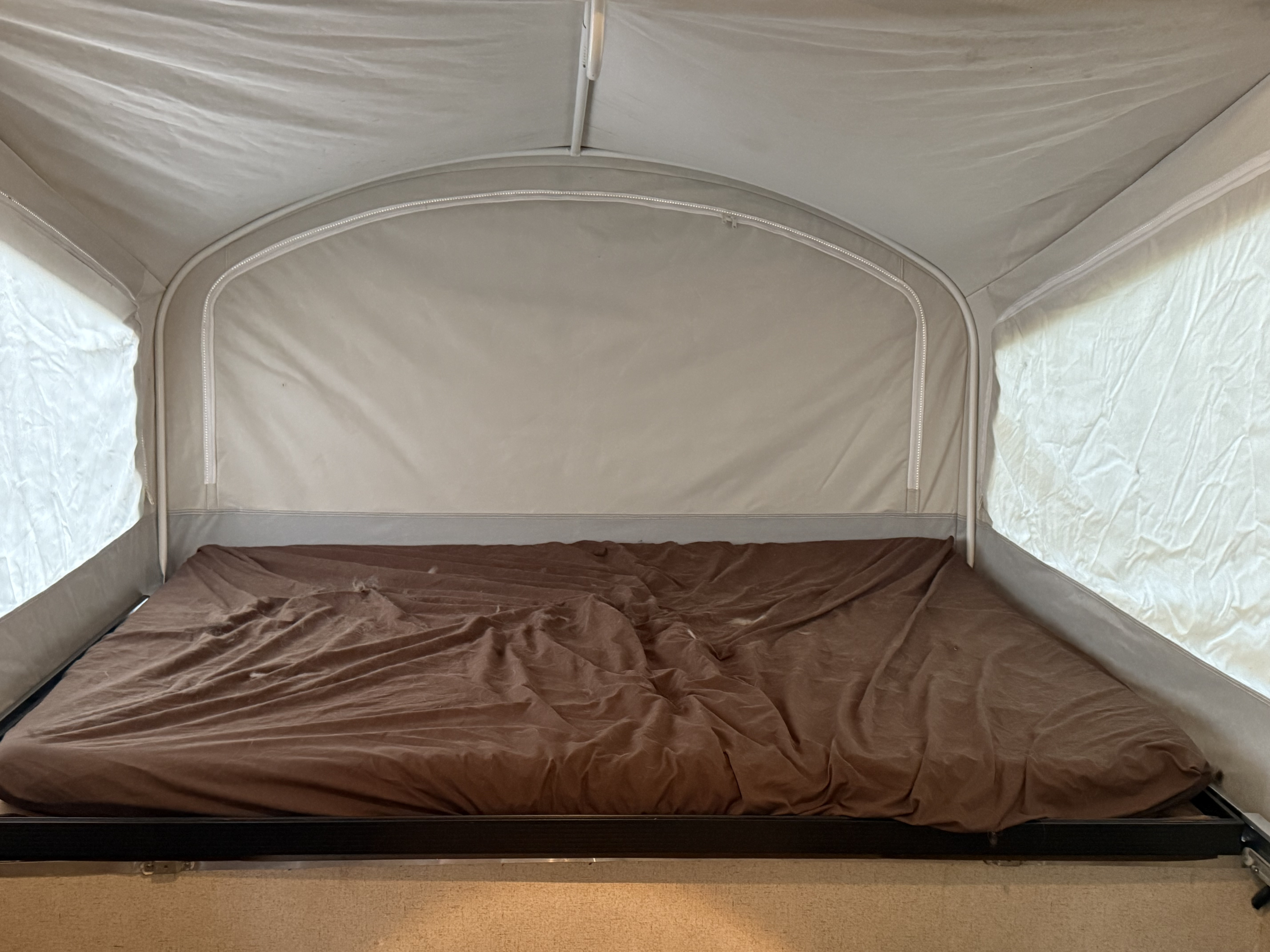 Inside an RV rental pop-up camper bed with rumpled brown sheets and light canvas tent walls. Zippers are visible on the fabric panels.. Jayco Jay 2016