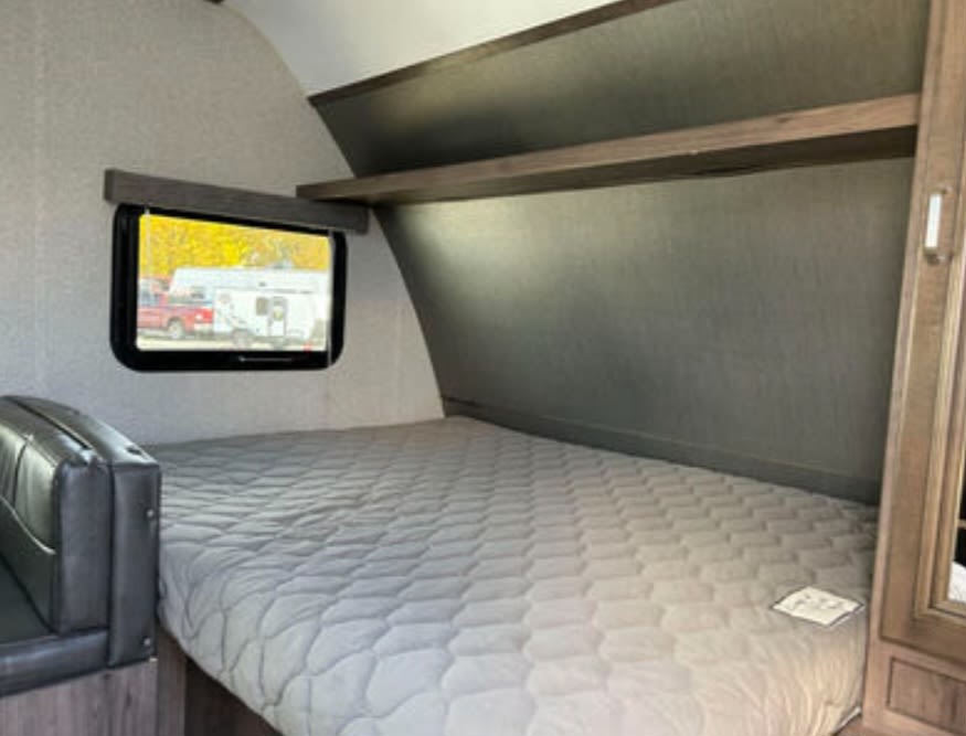 This RV rental bedroom features a cozy bed, window view, and shelving for a comfortable sleeping area on your trip.. Keystone RV Passport Express 2018
