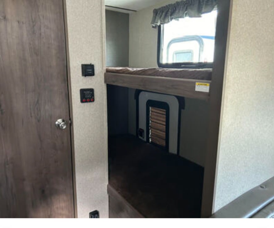 RV rental interior features compact bunk beds, a window with valance, and wall-mounted control panels beside a wooden door.. Keystone RV Passport Express 2018