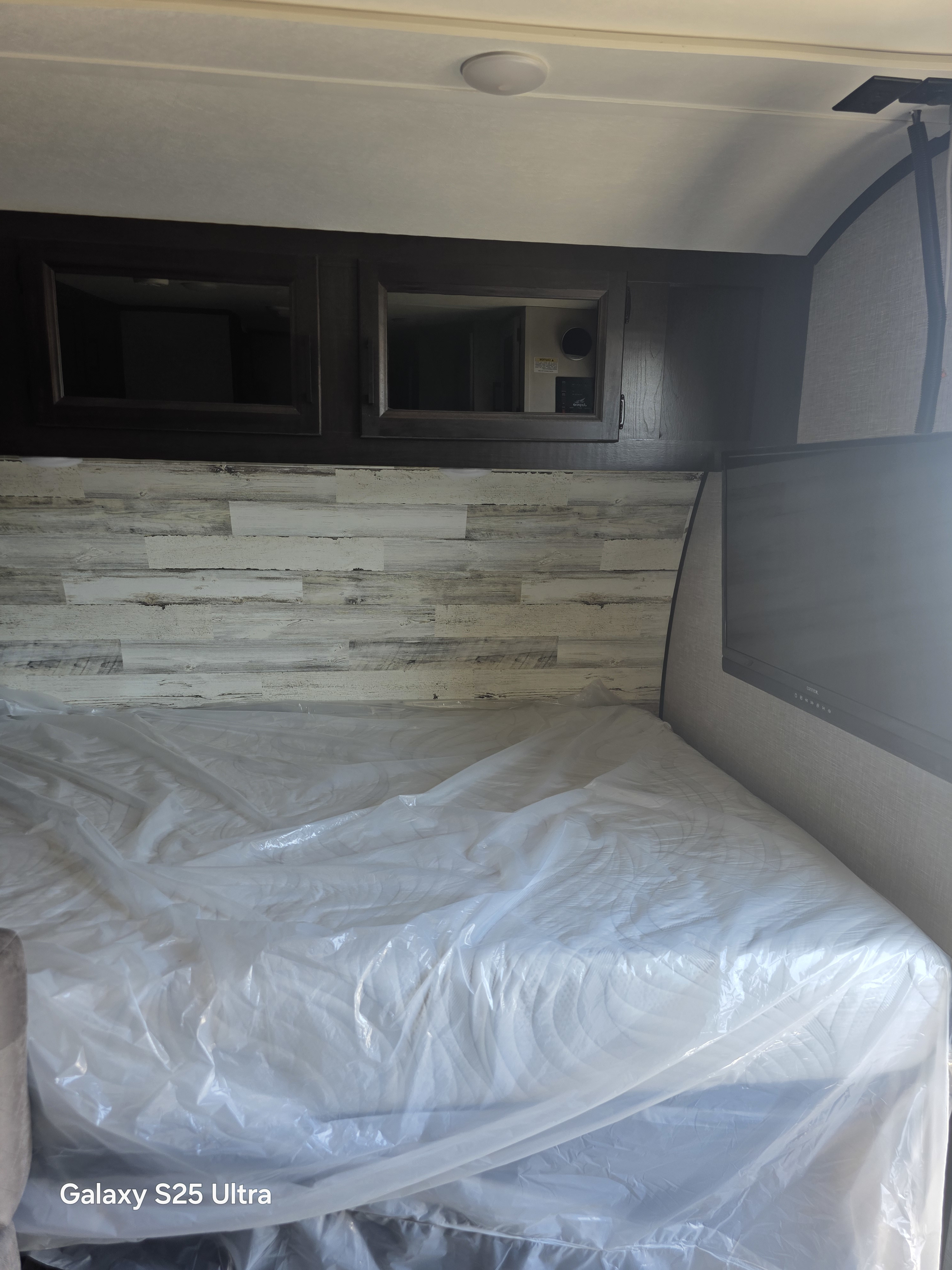 This RV rental bedroom features a new mattress in plastic, rustic wood-look headboard, overhead cabinets, TV, and ceiling light.. Jayco Jay Feather Micro 2023