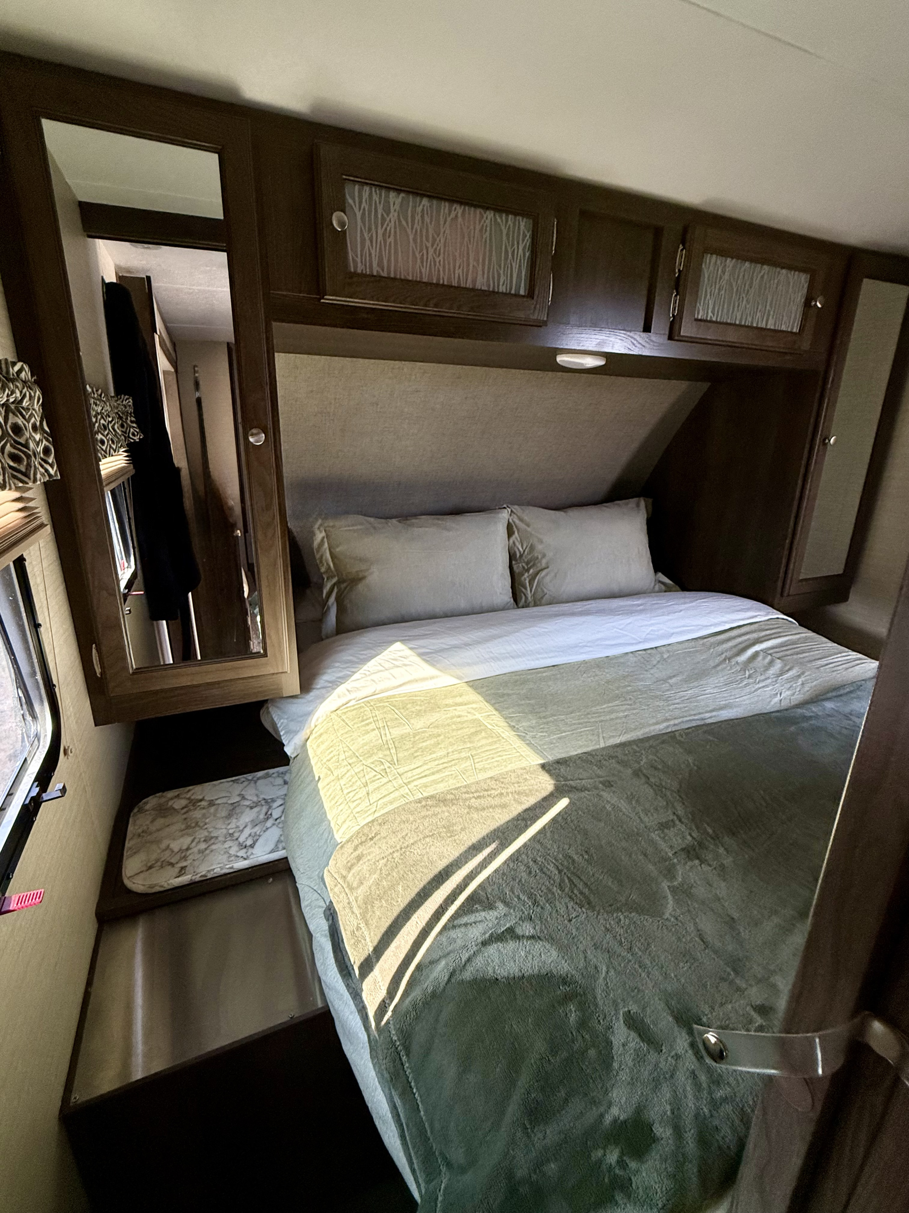 Inside a modern **rv rental**, a cozy bedroom featuring a sunlit bed, pillows, blankets, and dark wood overhead cabinets.. Keystone RV Bullet Ultra Lite 2018