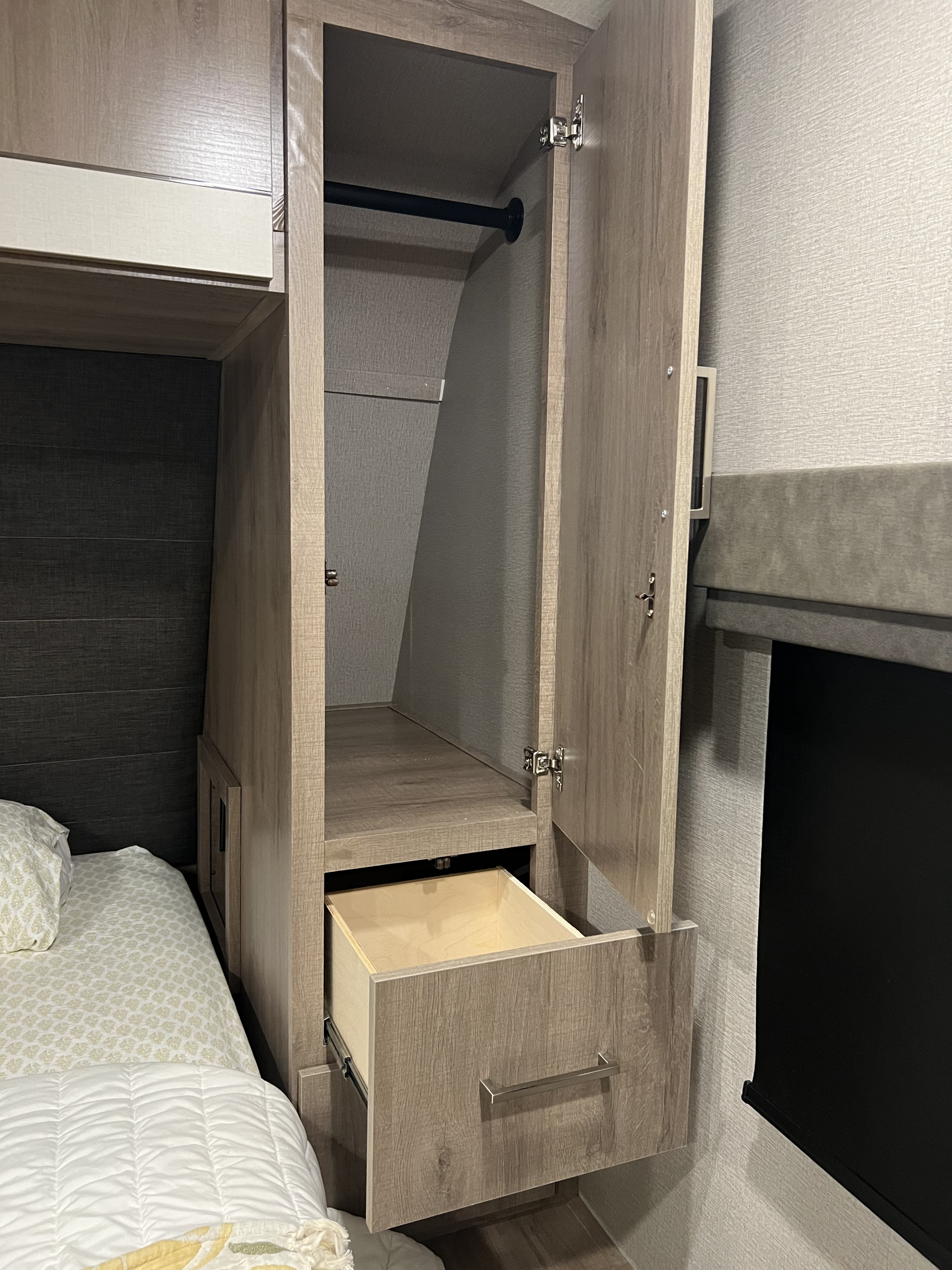 This RV rental shows a compact bedroom with an open wood-grain wardrobe, featuring a hanging rod, shelf, and open drawer, beside a bed.. Grand Design Imagine 2023