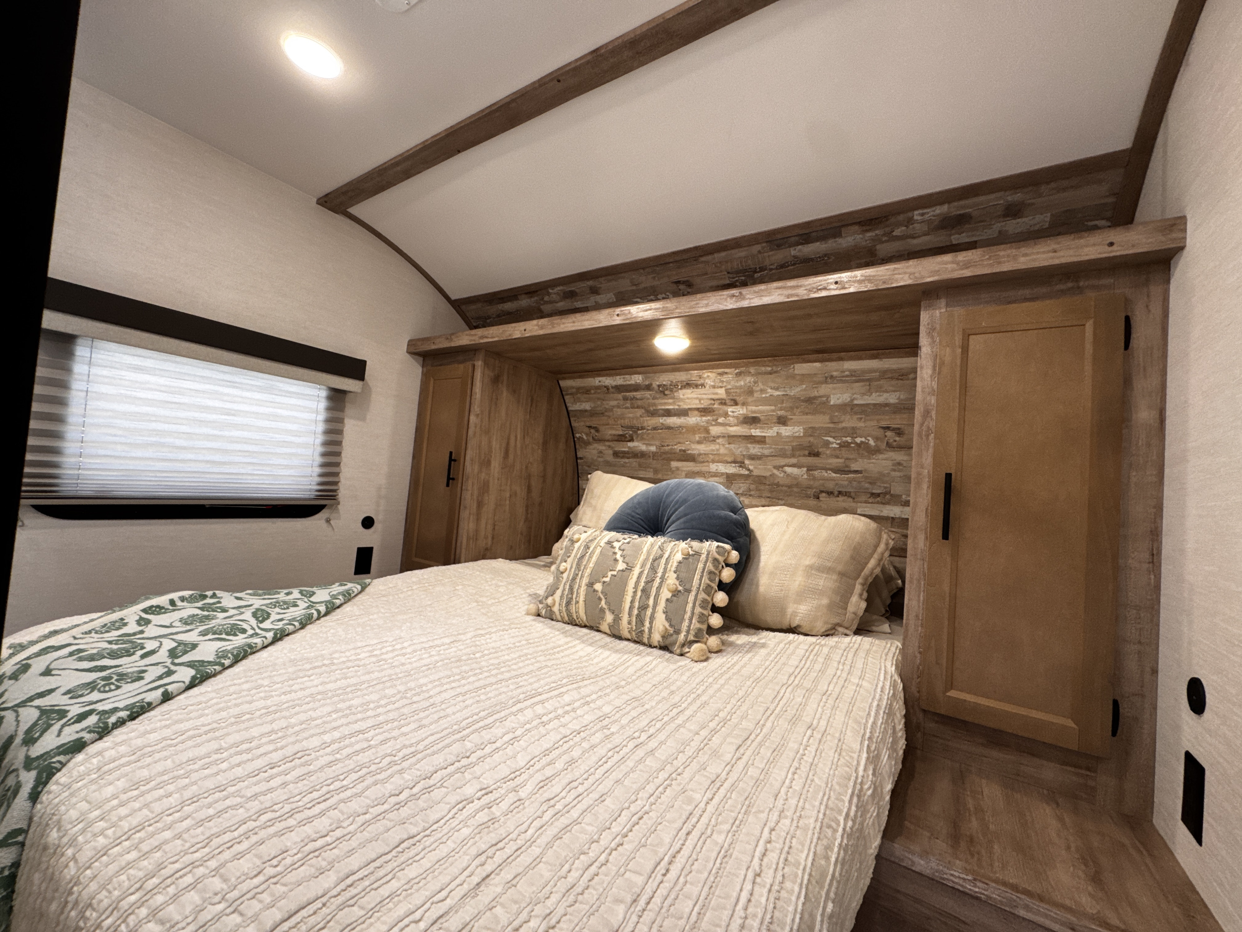 Inviting RV rental bedroom: bed with textured bedding, decorative pillows, window, and warm wood cabinetry.. Gulf Stream Enlighten 2025