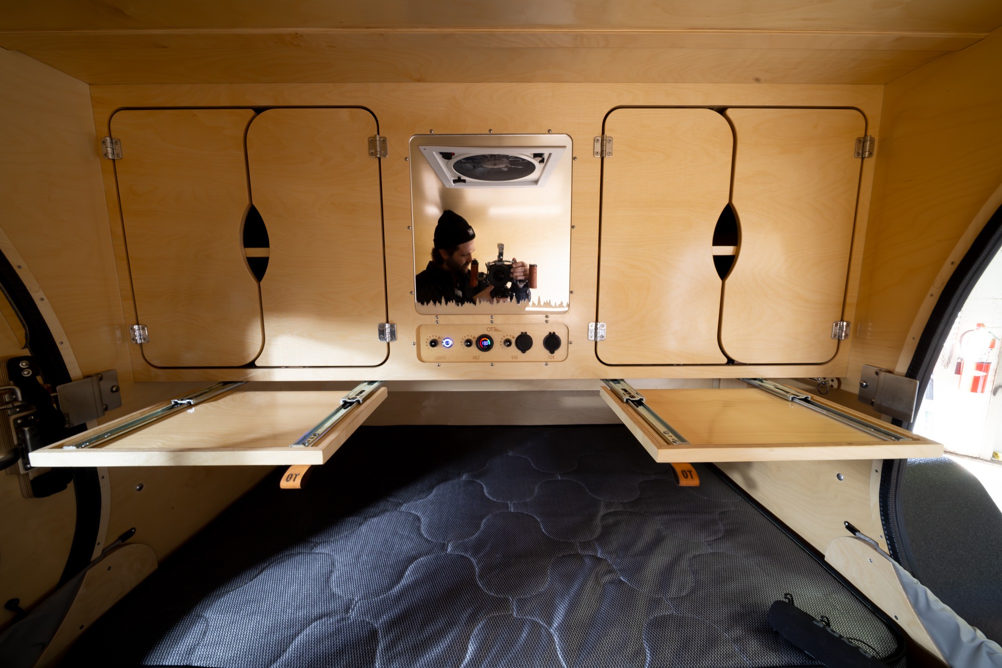 Teardrop RV rental interior: wooden cabinets, control panel with 12V/USB, pull-out tables, and photographer's mirror reflection.. Oregon Trail'r TerraDrop Alpha 2024